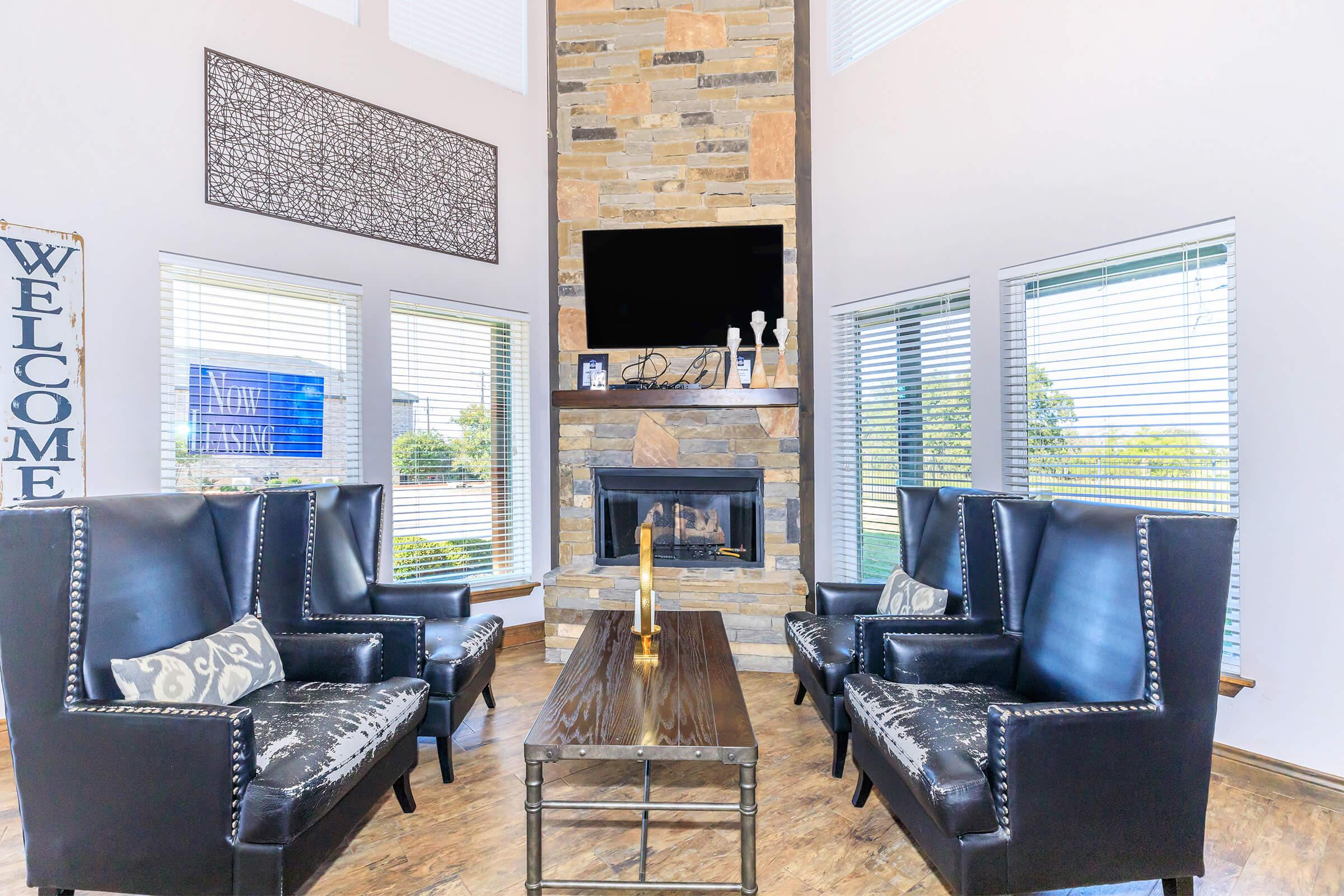 A spacious living room featuring two elegant black armchairs with decorative pillows, a wooden coffee table, and a modern stone fireplace. Large windows allow natural light to fill the space, and a flat-screen TV is mounted on the stone accent wall. A "Welcome" sign is visible on the left side.