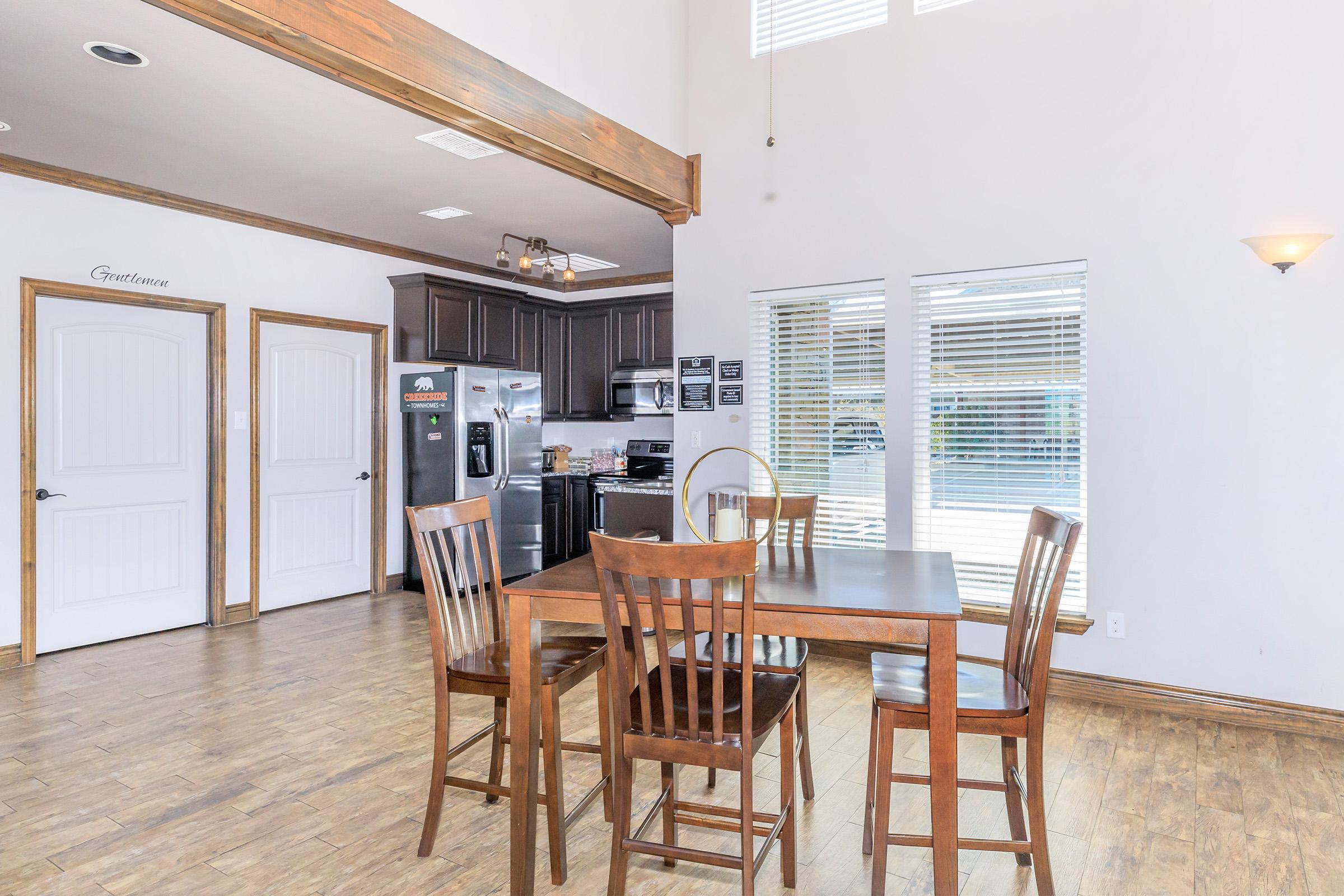 A bright dining area featuring a wooden table with four chairs, adjacent to a modern kitchen with stainless steel appliances. Large windows allow natural light to fill the space, highlighting the light-colored walls and wooden flooring. The room has a cozy, inviting atmosphere.