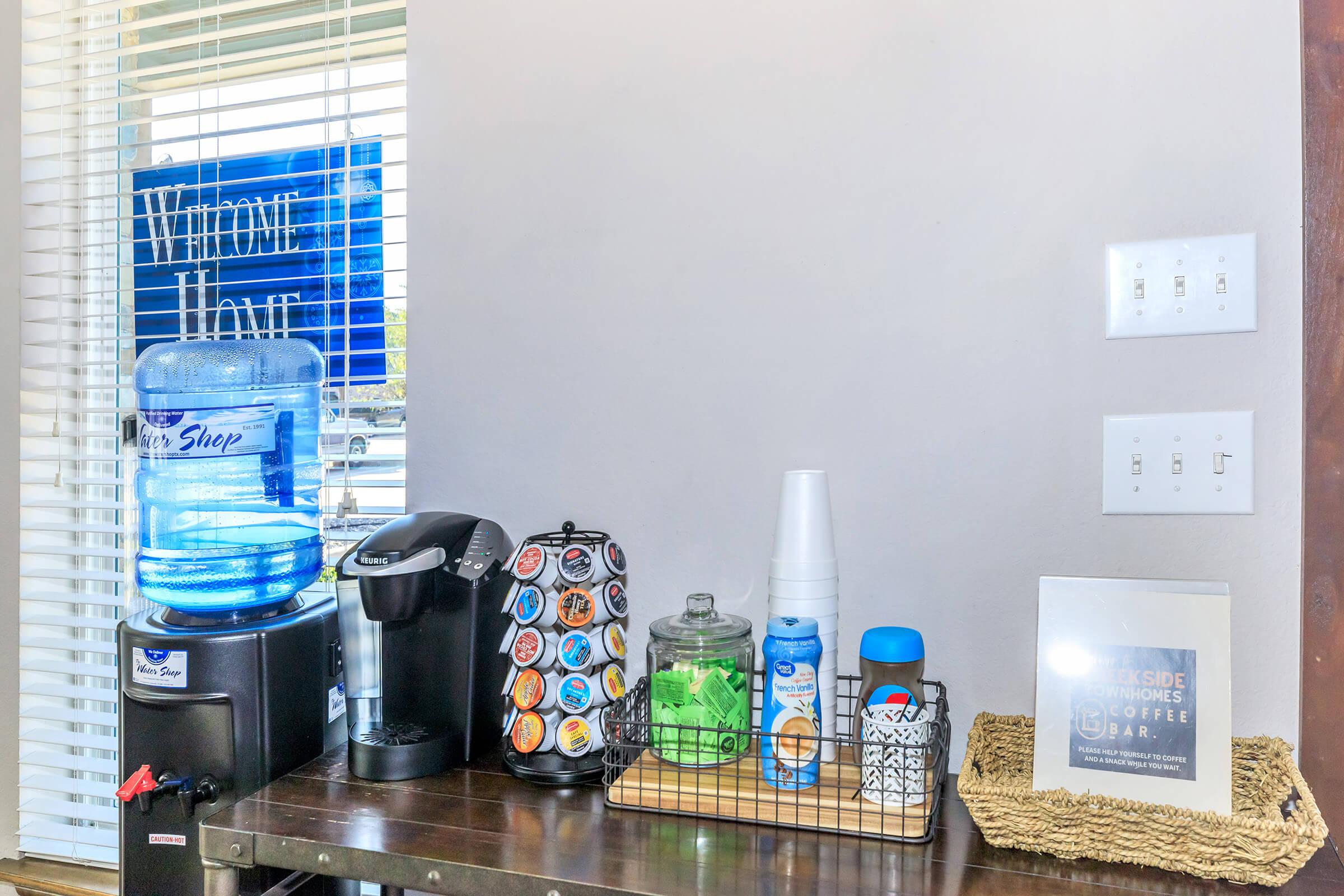 A well-set coffee station featuring a blue water dispenser, a coffee maker with colorful coffee pods, a glass jar, and a white cup. Thereβs a decorative sign stating "Welcome Home" on the wall above the station, and light-colored walls with a window allowing natural light in.