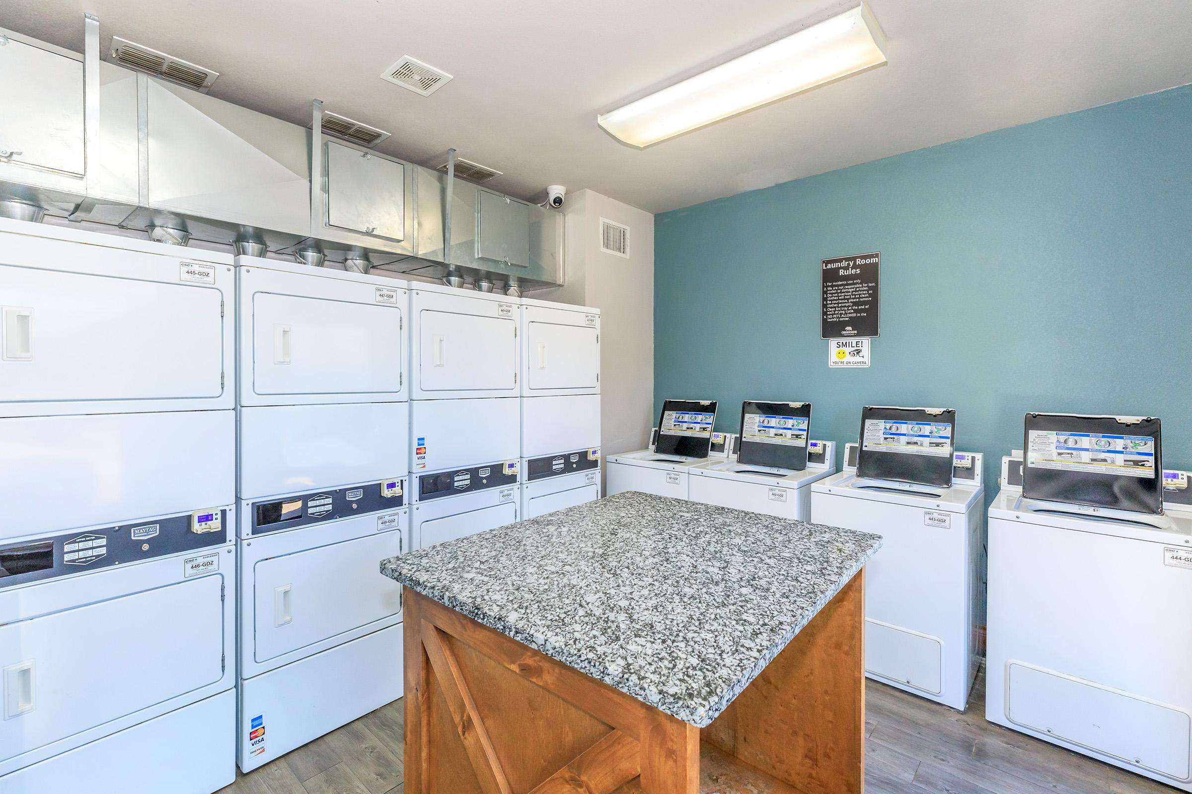 Laundry room featuring multiple white washing machines and dryers arranged against one wall. There is a granite countertop in the center of the room. The walls are painted a calming blue, with a bulletin board displaying information. Bright overhead lighting illuminates the space.