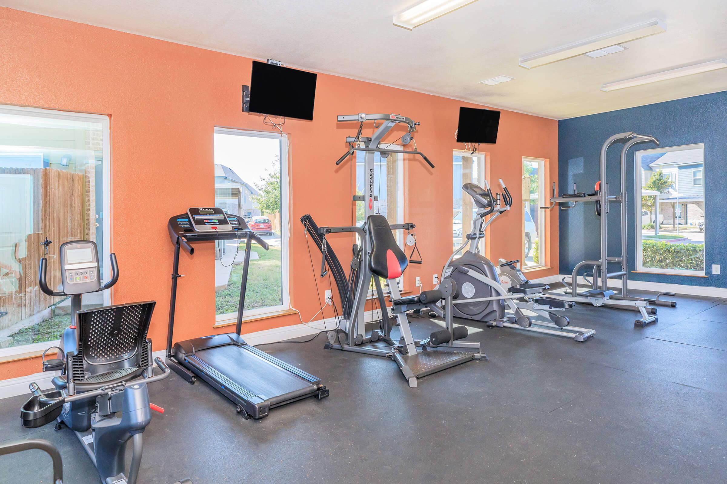 A bright and spacious gym featuring a variety of exercise equipment, including a treadmill, elliptical machine, and weight station. Large windows provide natural light and an inviting atmosphere. Two flat-screen TVs are mounted on the orange wall, enhancing the workout experience.