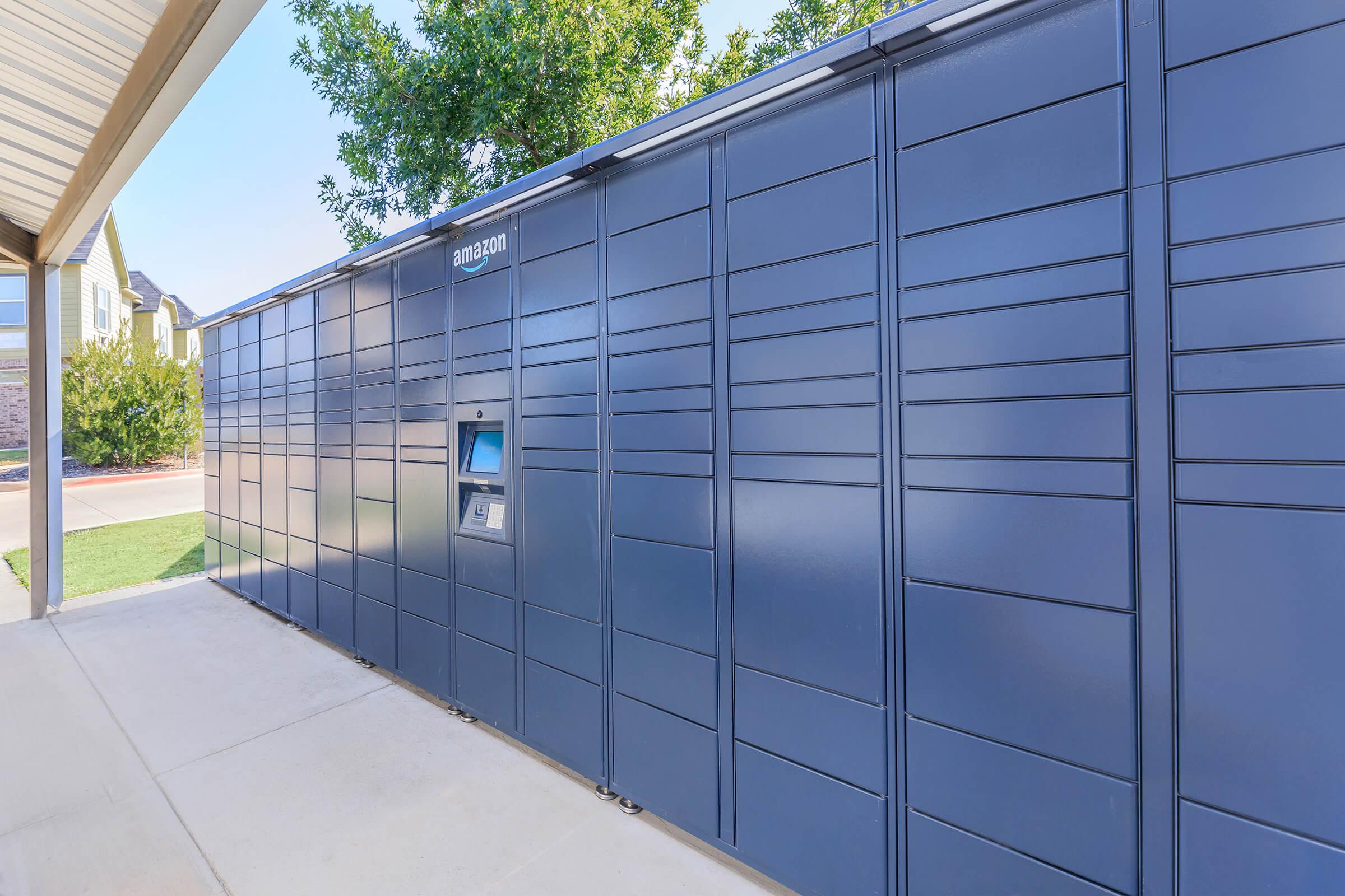 A row of blue Amazon lockers installed outside in a residential area, providing secure package delivery and pickup options. Green grass and a clear sky are visible in the background.