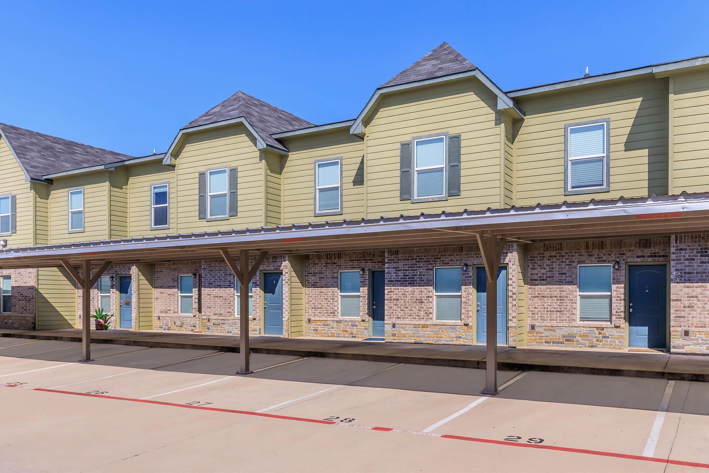 Row of two-story townhouse units with a greenish-yellow exterior and brick accents. Each unit features a covered parking area below, with numbered spaces clearly visible on the ground. The sky is clear and blue, enhancing the overall bright appearance of the property.