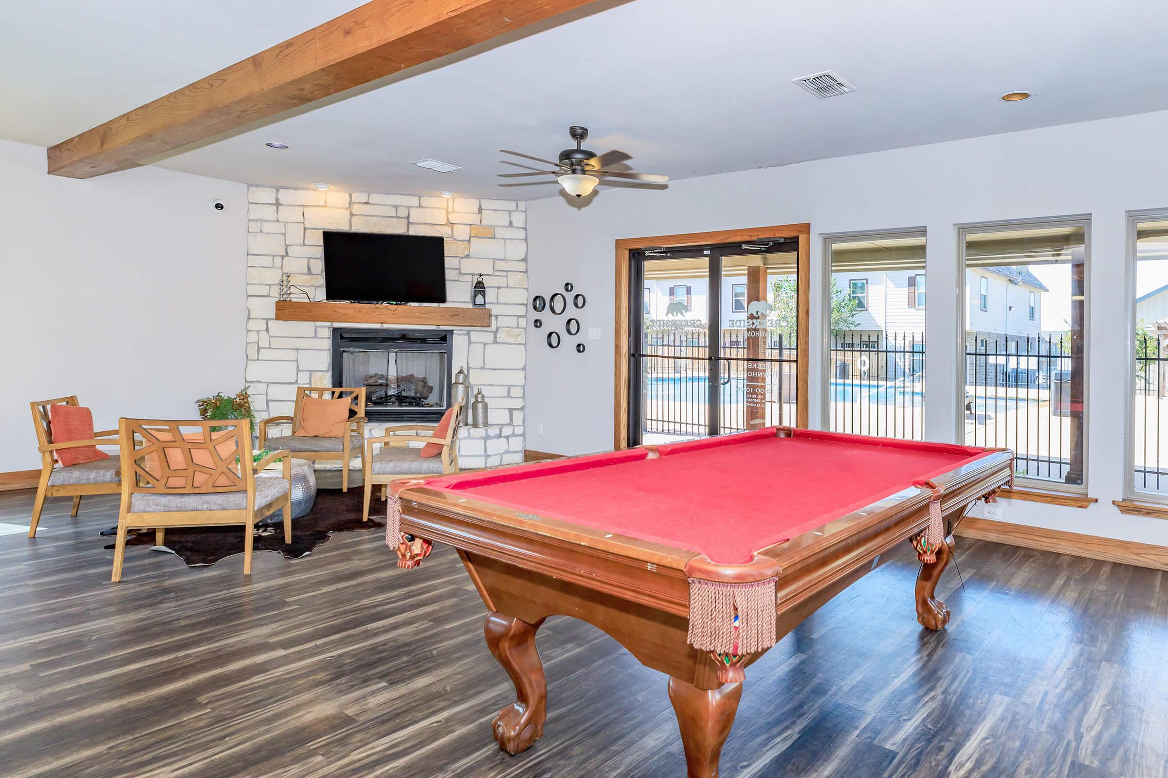 Interior view of a modern game room featuring a red felt pool table in the center. A stone fireplace and a flat-screen TV are on one wall, while large windows overlook an outdoor area. Comfortable seating with decorative chairs enhances the inviting atmosphere.