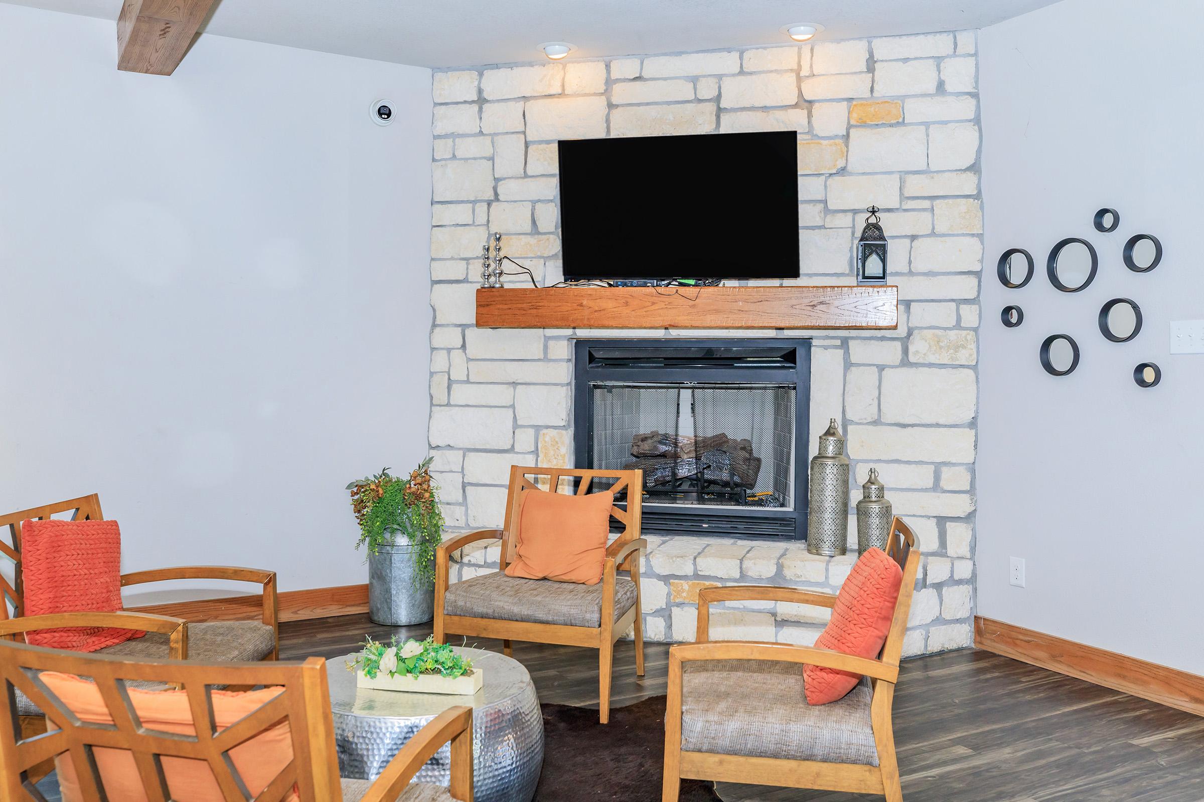 A cozy living area featuring a stone fireplace with a mounted TV above it. Four wooden chairs with orange cushions are arranged around a small table. The room has a modern aesthetic with a neutral color palette and decorative elements like lanterns and circular wall art.