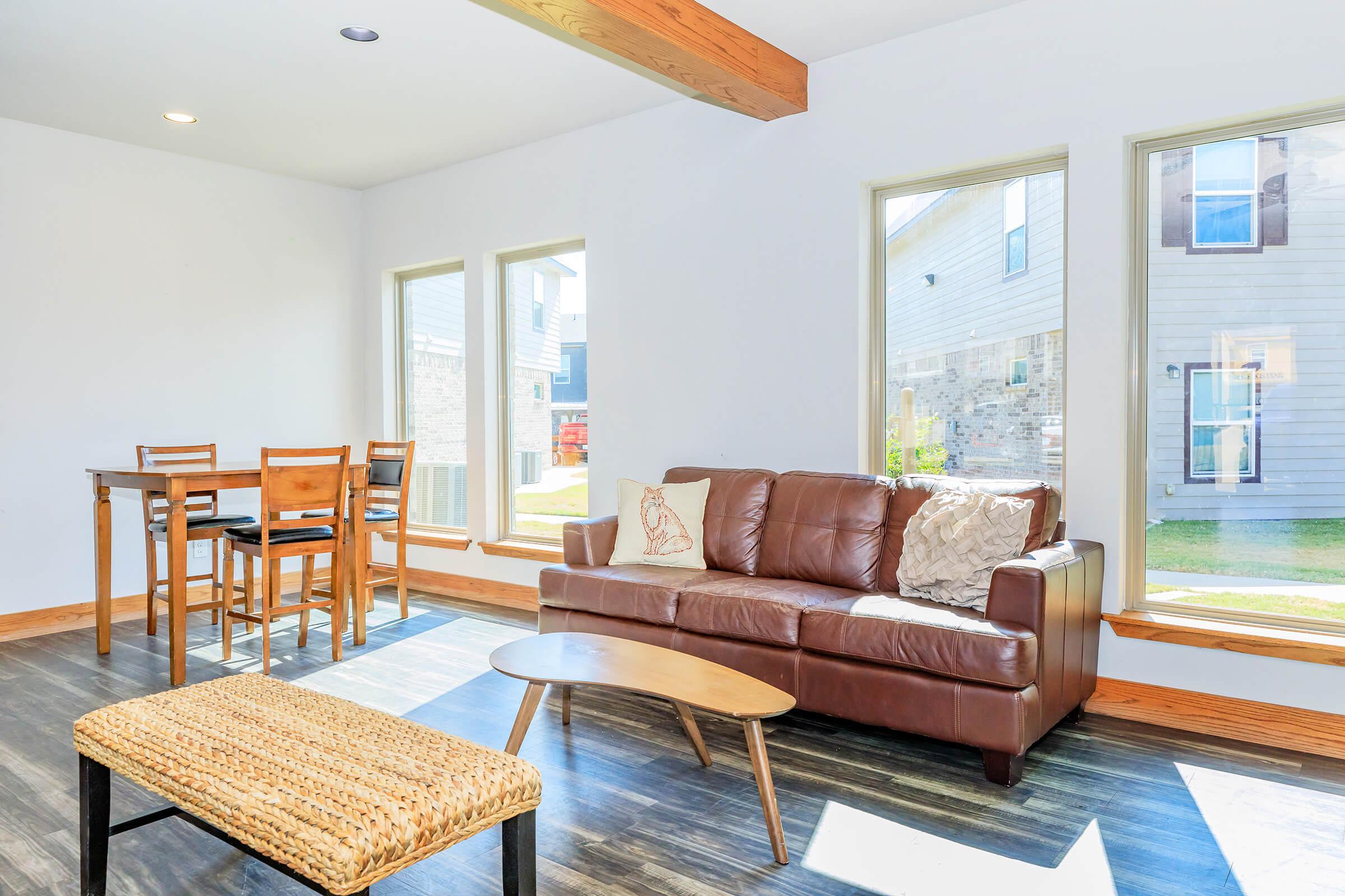 Bright and modern living space featuring a brown leather sofa with decorative pillows, a light wooden coffee table, and a woven ottoman. Natural light floods the room through large windows. A dining set with wooden chairs is visible in the background, enhancing the cozy ambiance.