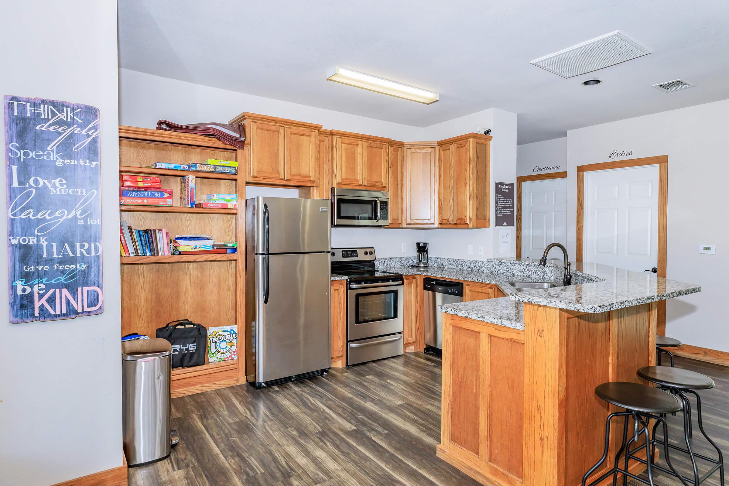 A modern kitchen with wooden cabinets and stainless steel appliances, including a refrigerator, oven, and microwave. The countertop is made of granite, and there are bar stools next to the counter. Shelves display books and kitchen items, and a motivational quote is framed on the wall.