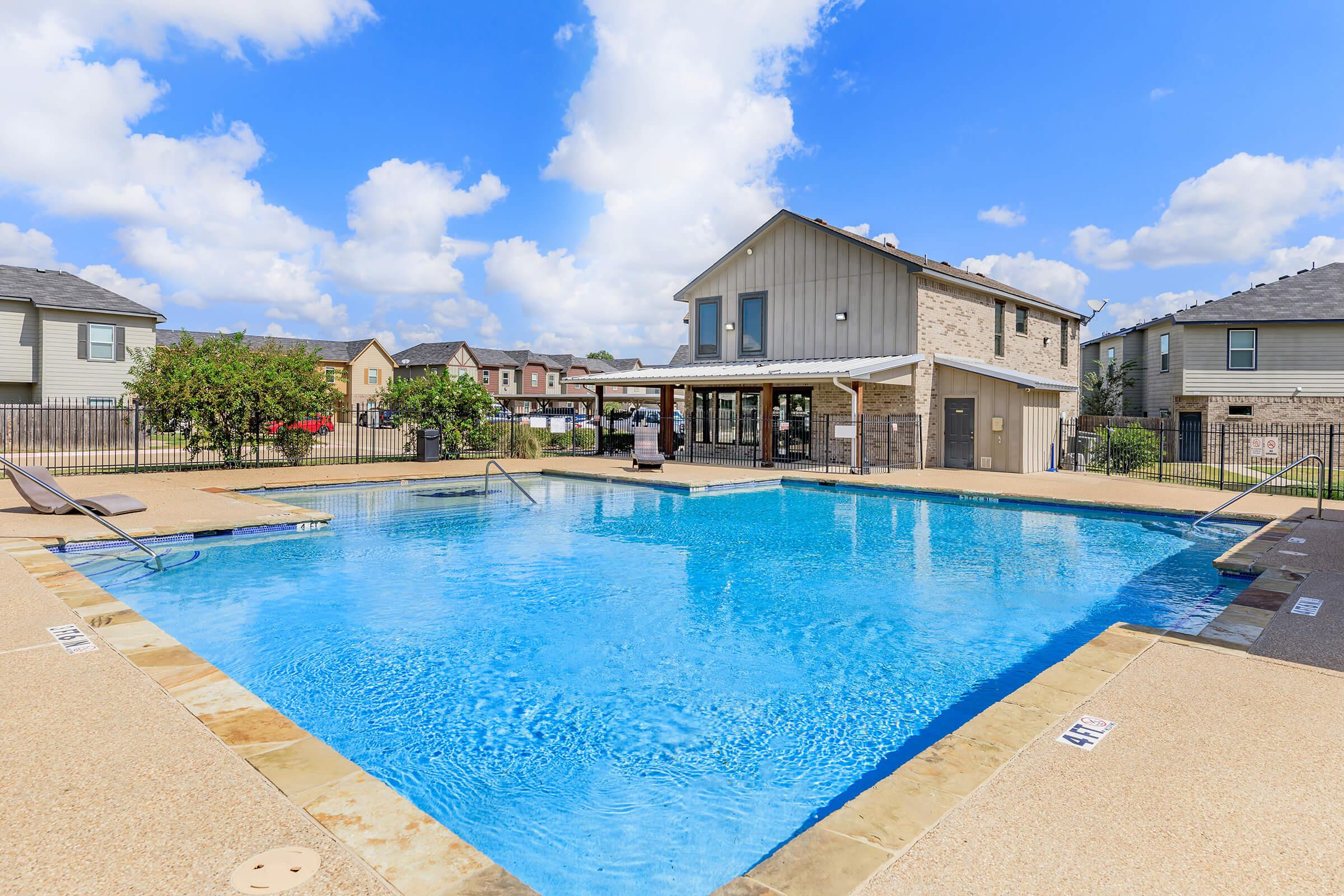 A sunny outdoor swimming pool surrounded by tan and stone decking. The pool area includes lounge chairs and is bordered by a fence. In the background, there are several residential buildings, and fluffy clouds are visible in a bright blue sky.