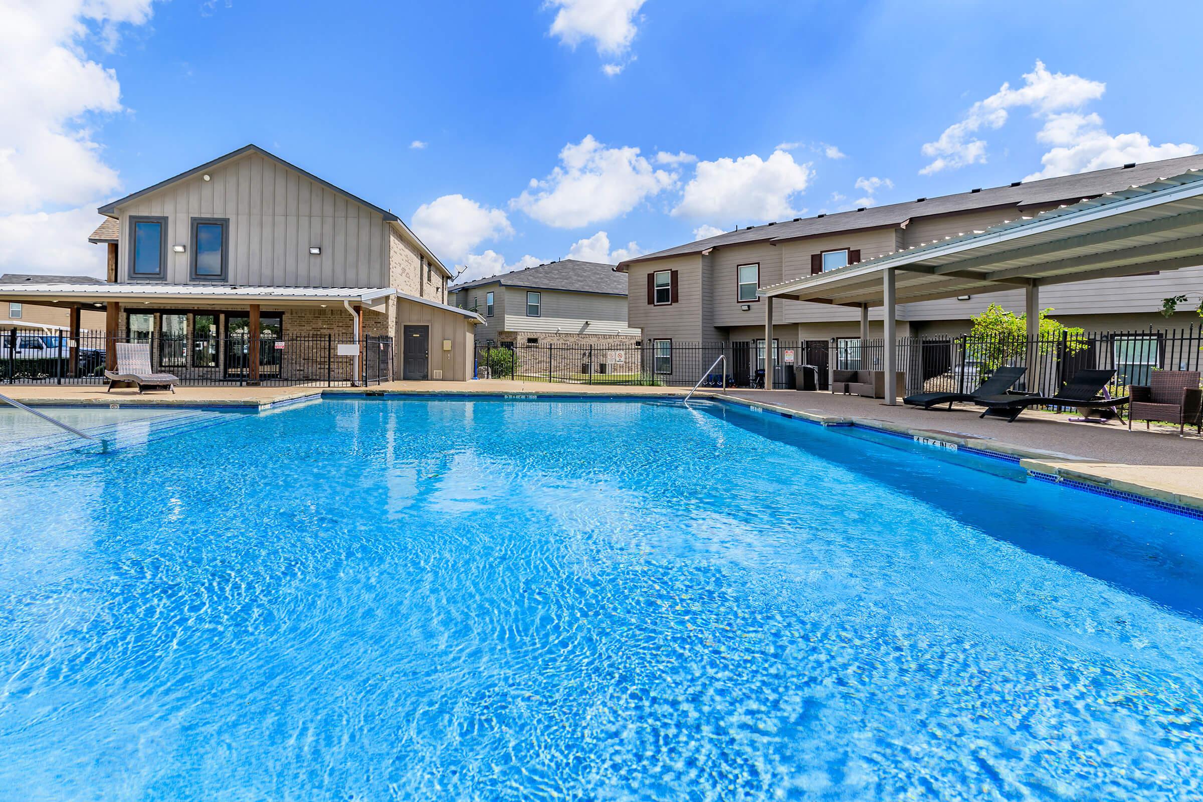A clear blue swimming pool surrounded by lounge chairs, with a two-story building on one side and additional residential buildings in the background. The sky is bright with a few fluffy clouds, creating a welcoming atmosphere for relaxation and recreation.