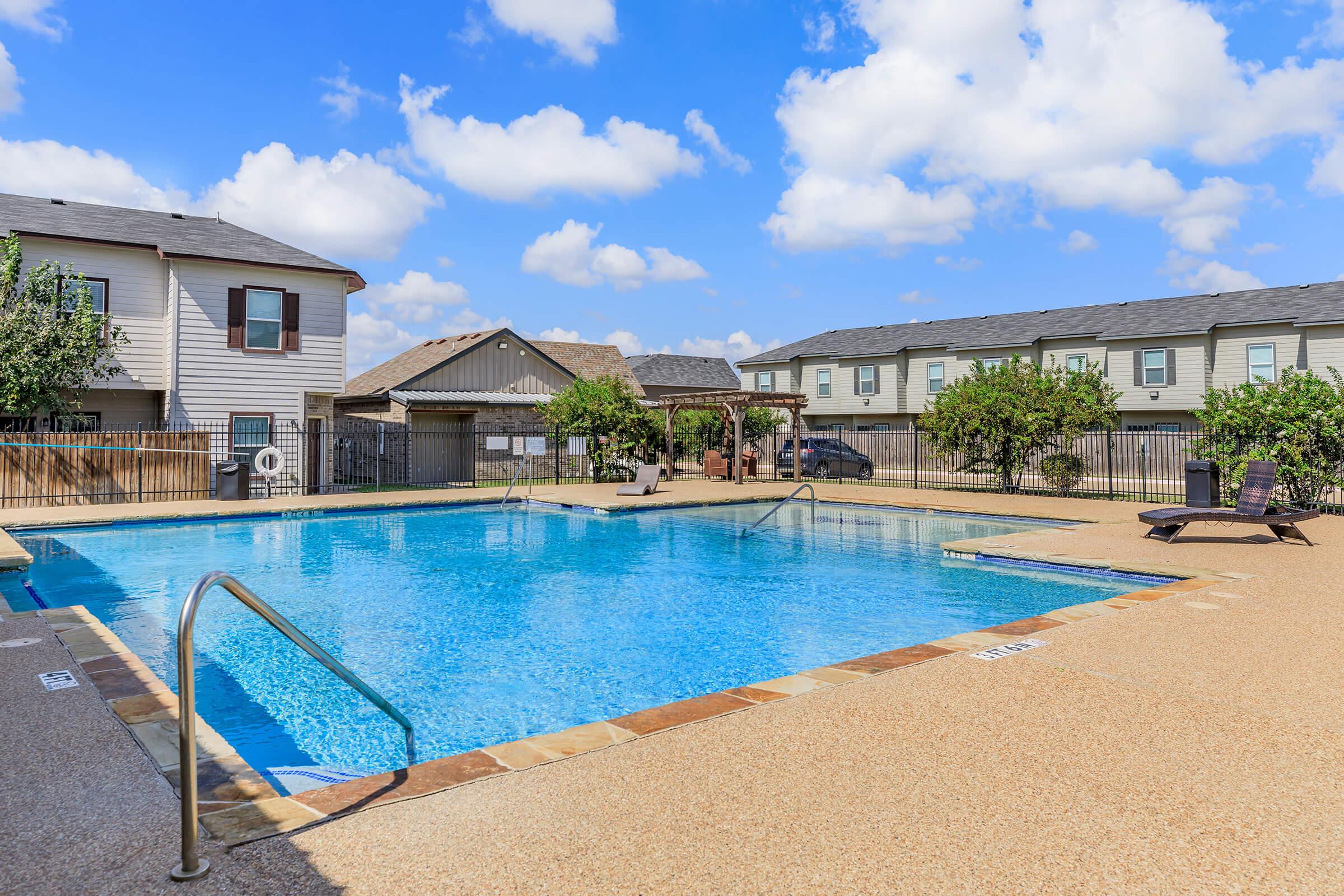 A clean swimming pool surrounded by a patio area, with lounge chairs and a shaded gazebo. In the background, there are residential buildings and clear blue skies with fluffy clouds.