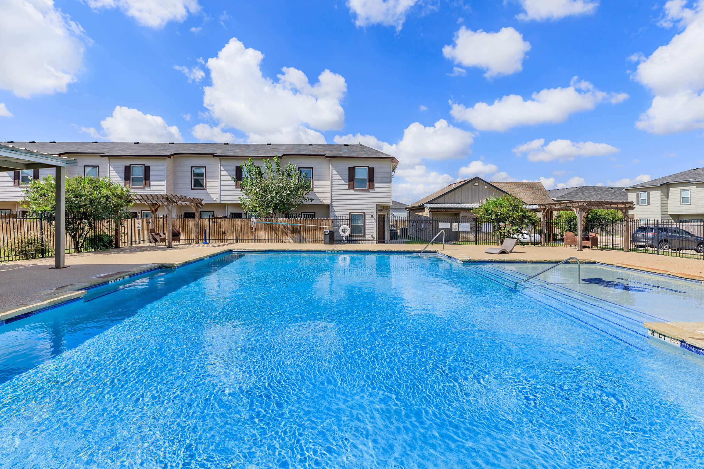 A sparkling blue swimming pool surrounded by lounge chairs and greenery, with two buildings in the background. The sky is bright and partly cloudy, creating a cheerful atmosphere. The pool area is well-maintained, offering a relaxing outdoor space for residents or guests.