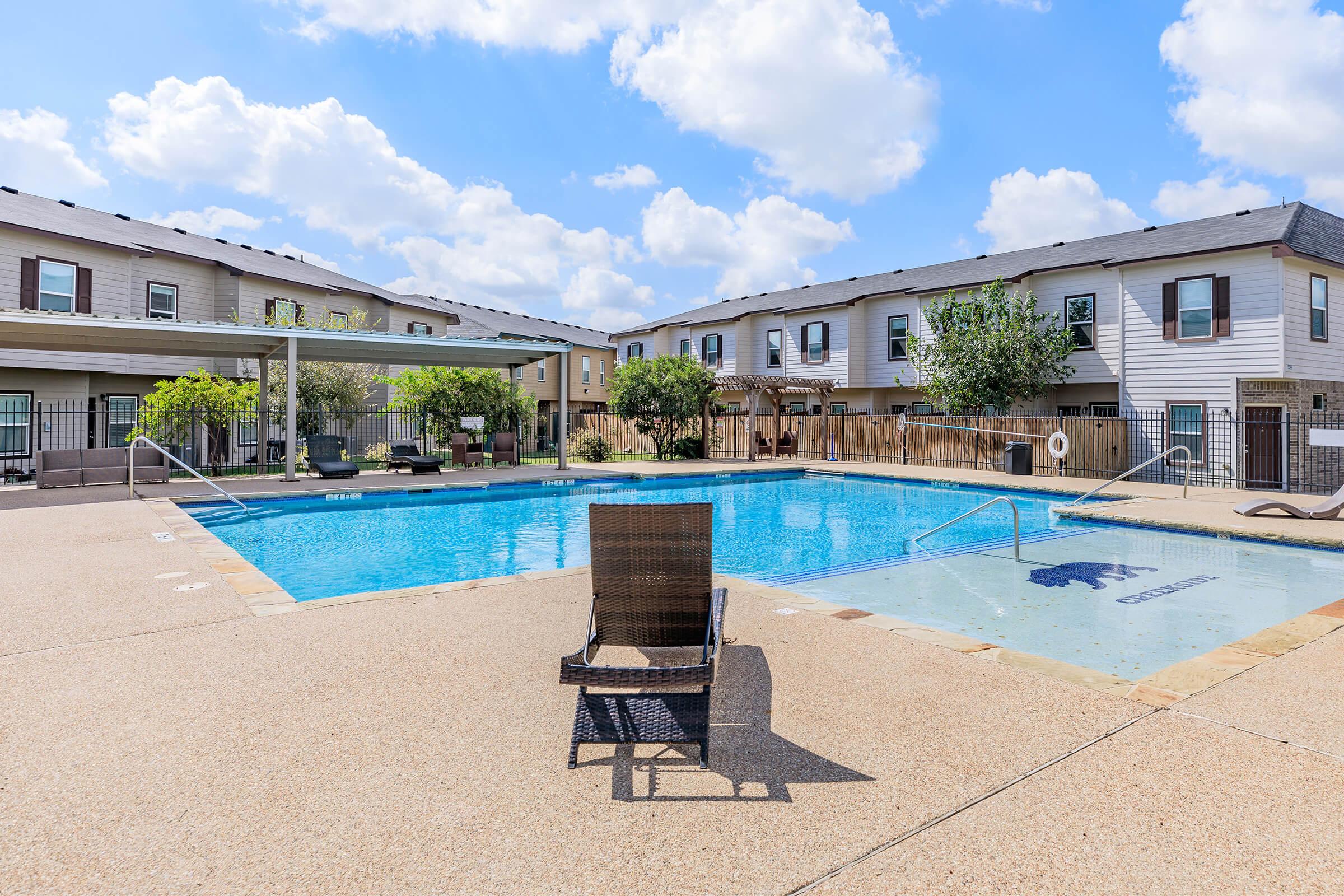 A sunny outdoor area featuring a clear blue swimming pool surrounded by lounge chairs and greenery. In the background, there are two-story residential buildings with multiple windows and a shaded patio area. The sky is bright with fluffy clouds.