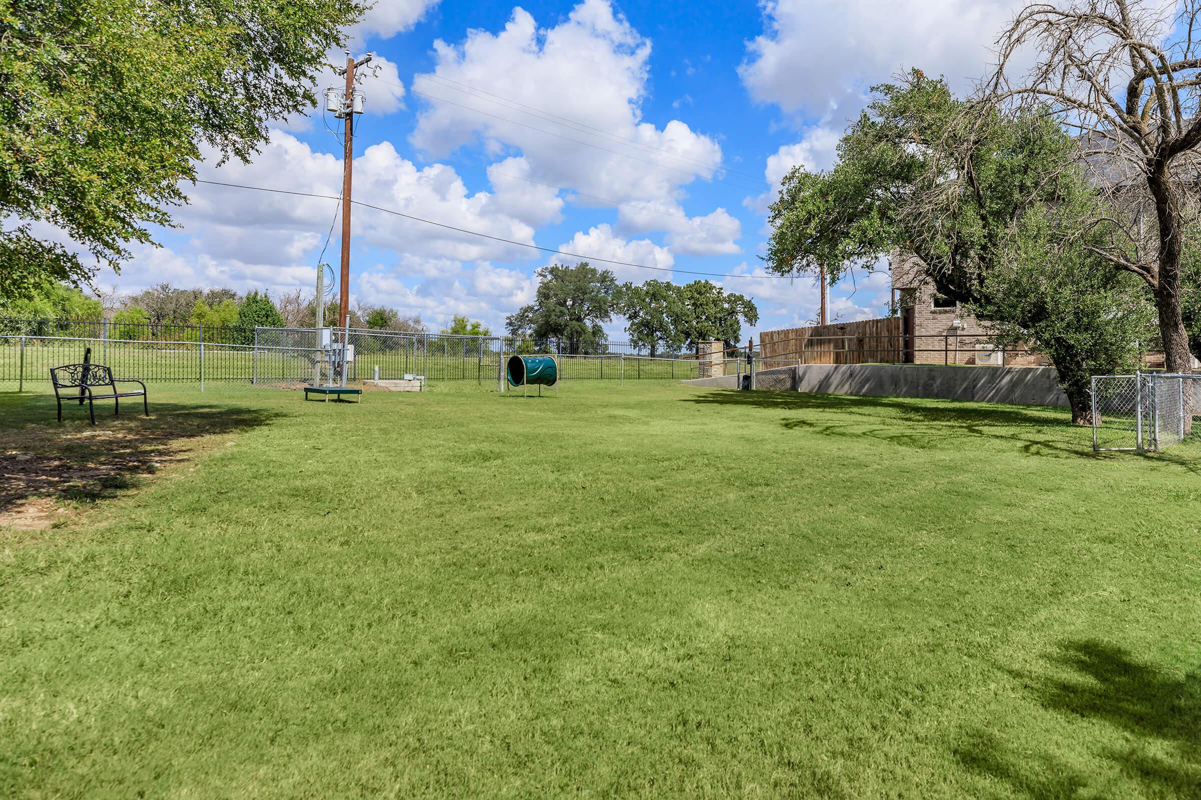 A spacious grassy area in a park featuring a chain-link fence, a bench, a covered structure, and a cylindrical play feature. The sky is bright with fluffy white clouds, and trees line the background, creating a tranquil outdoor environment.
