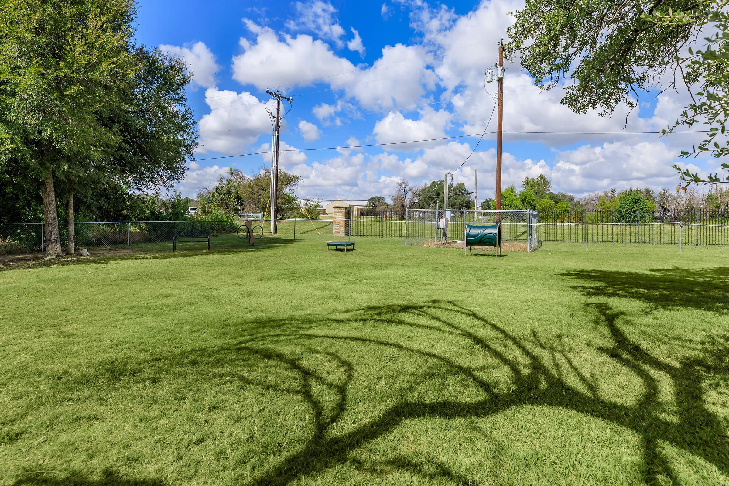 A spacious green park with well-maintained grass. In the background, there are a few trees and a fenced area. A picnic table is visible on the grass, along with a large trash bin. The sky is partly cloudy with blue skies and scattered clouds, creating a pleasant outdoor atmosphere.