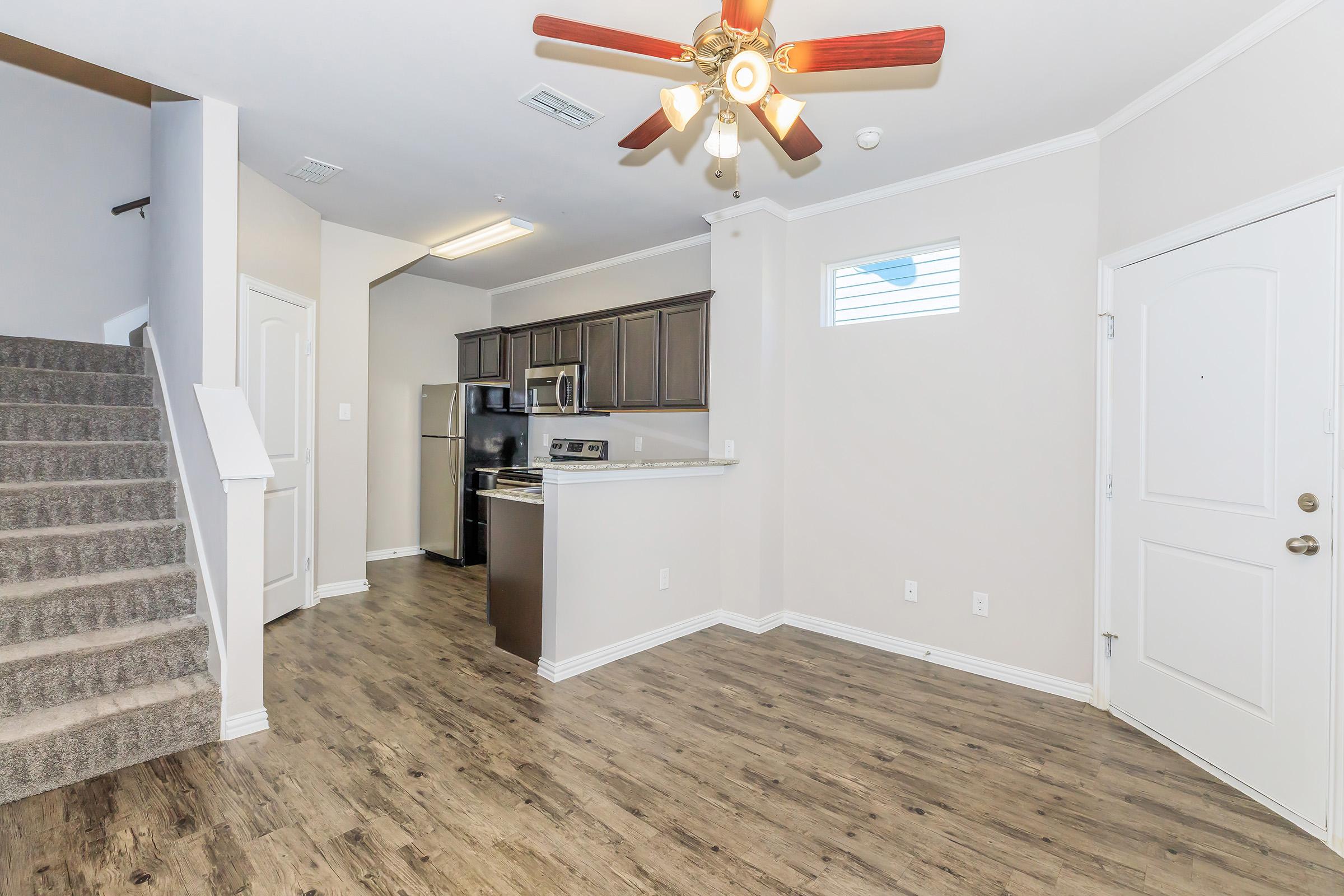 A view of a modern living space featuring a staircase, kitchen area with dark wood cabinets and stainless steel appliances, and a door leading outside. The floor is laminate wood, and there is a ceiling fan above. Natural light enters through a window, creating a bright atmosphere.