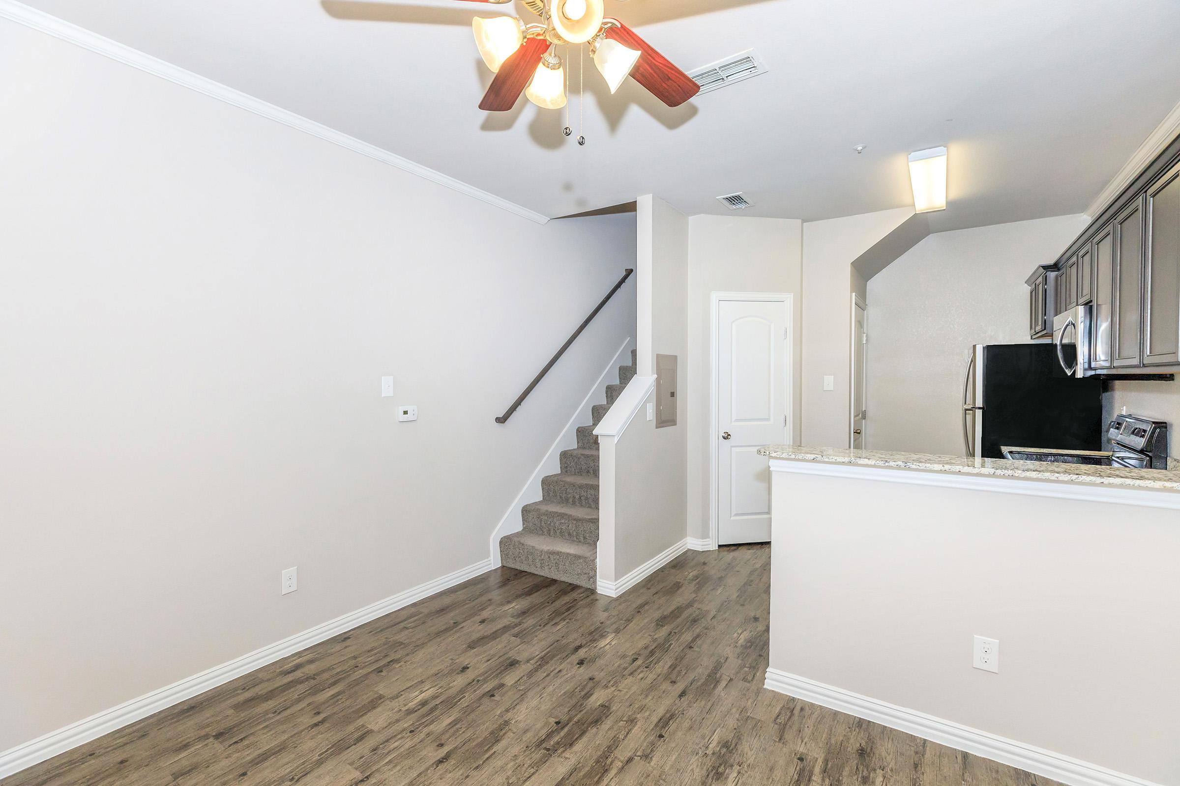 Interior view of a modern apartment featuring a living area with wood-like flooring, a ceiling fan, and a staircase leading to an upper level. The kitchen includes granite countertops and stainless steel appliances. Neutral wall colors create a bright and airy atmosphere.