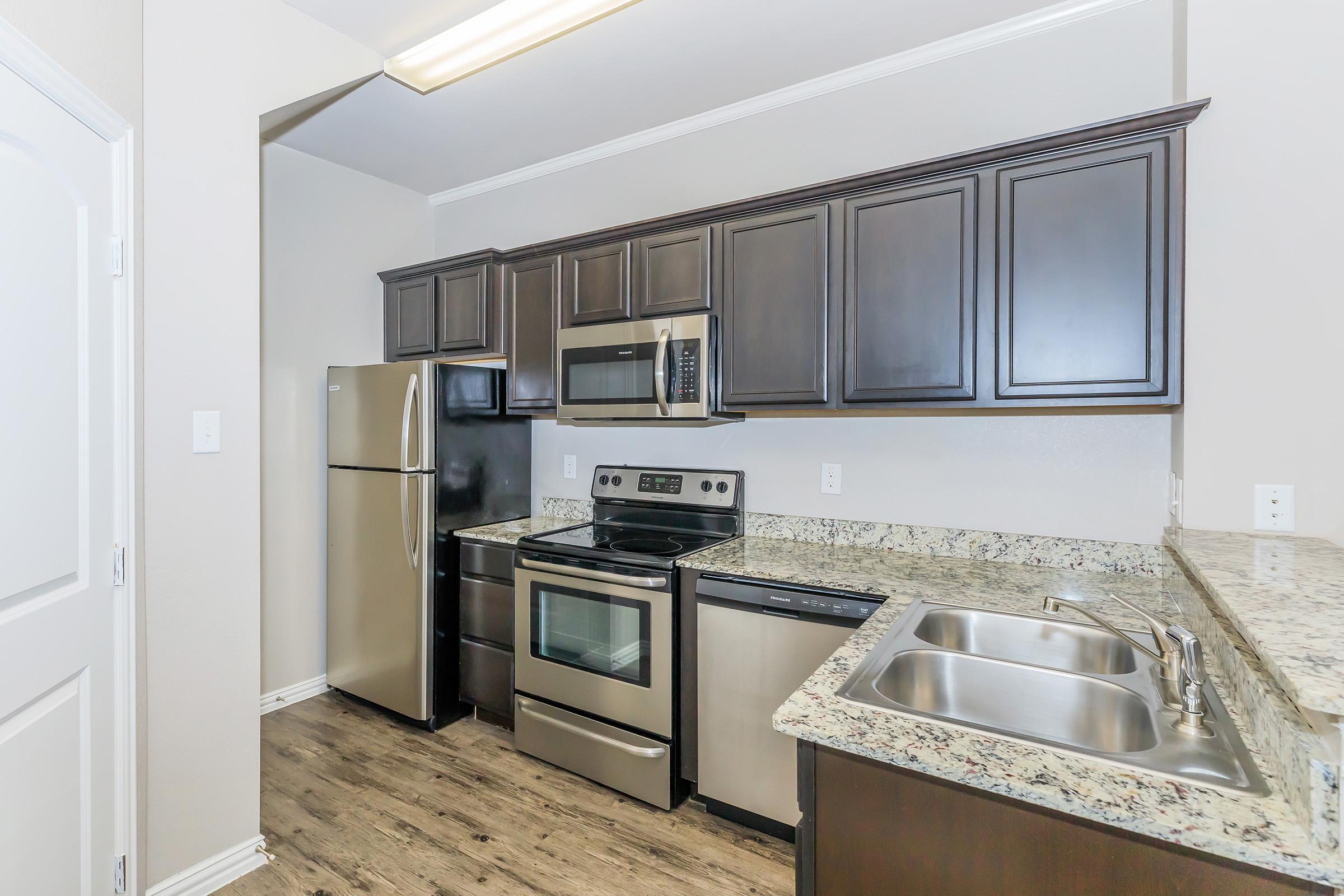 Modern kitchen featuring dark wooden cabinets, stainless steel appliances including a refrigerator, microwave, oven, and dishwasher. The countertop is made of speckled granite, and there is a double sink. The space has a clean, contemporary design with laminate flooring.