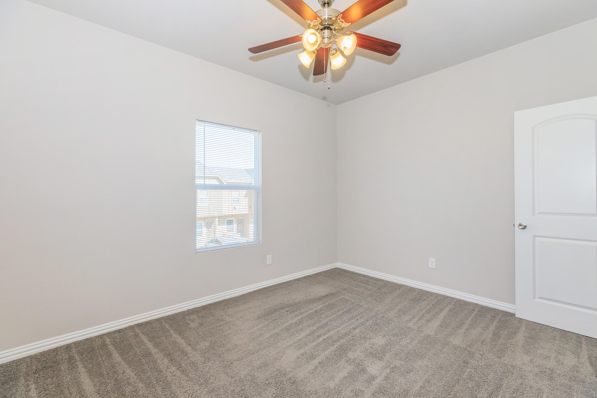 Empty room with light gray walls and carpeted flooring, featuring a ceiling fan with three light bulbs. A window allows natural light to enter, and there is a white door on one side. The room is unadorned, creating a clean and spacious atmosphere.