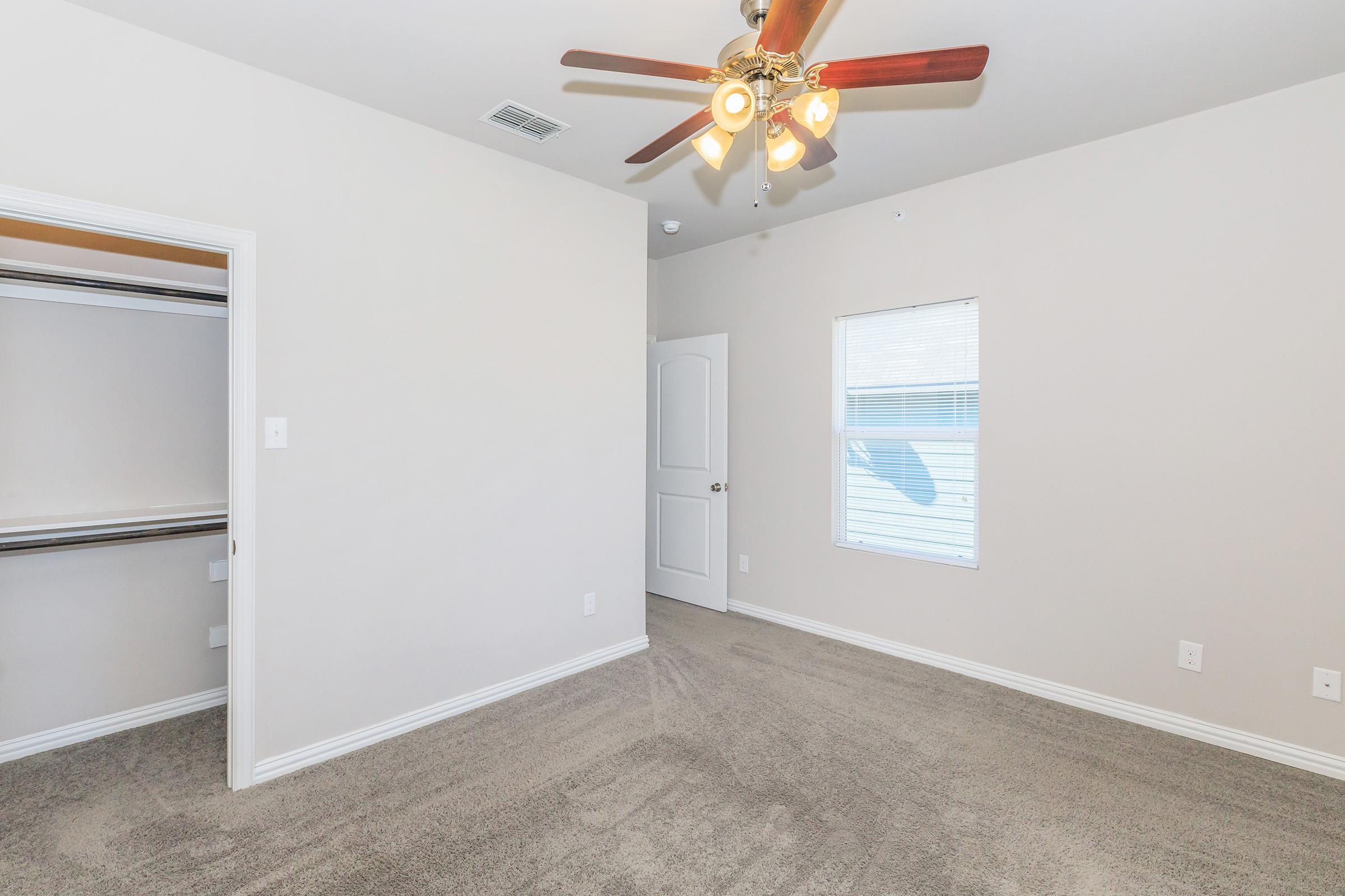 A well-lit, empty bedroom featuring beige walls and carpet, a ceiling fan with lights, and a closet with sliding doors. A white door leads to another room, and there's a window allowing natural light to enter. The overall space is clean, inviting, and ready for furnishings.