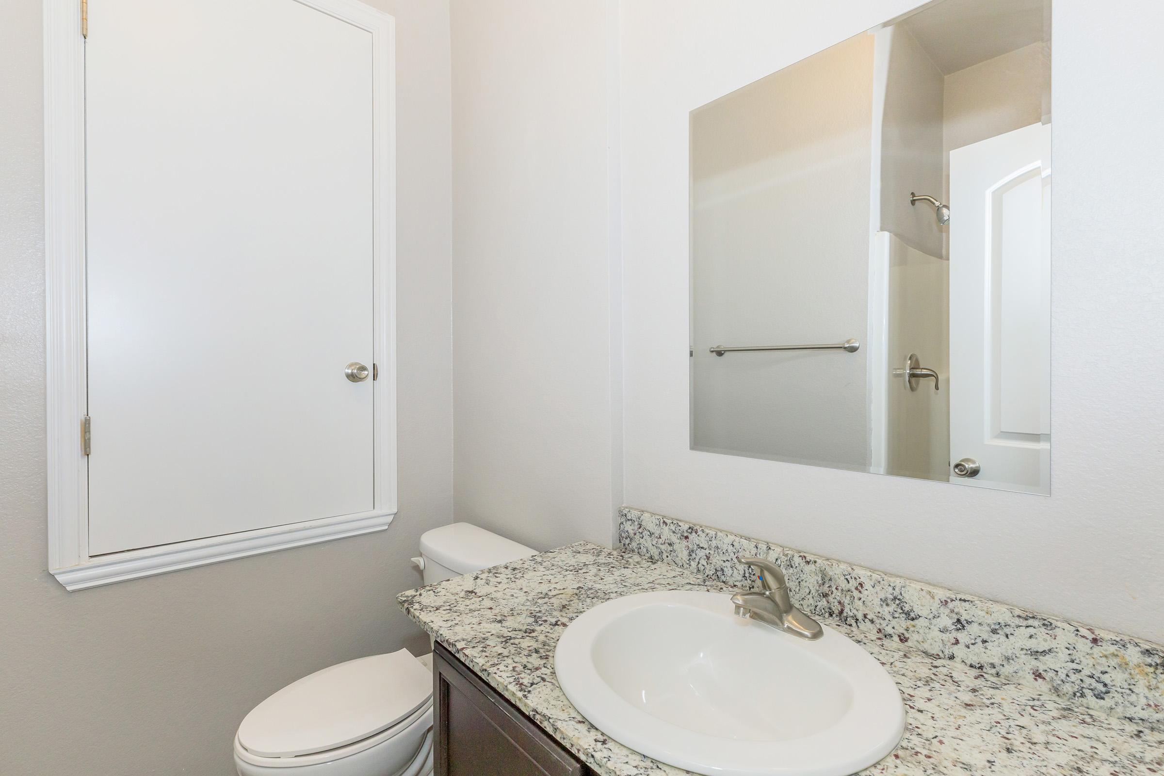 A modern bathroom featuring a white toilet, a sink with a granite countertop, and a mirror above the sink. The walls are painted in a light color, and there is a door leading to a shower area, which is partially visible. The overall design is clean and contemporary.