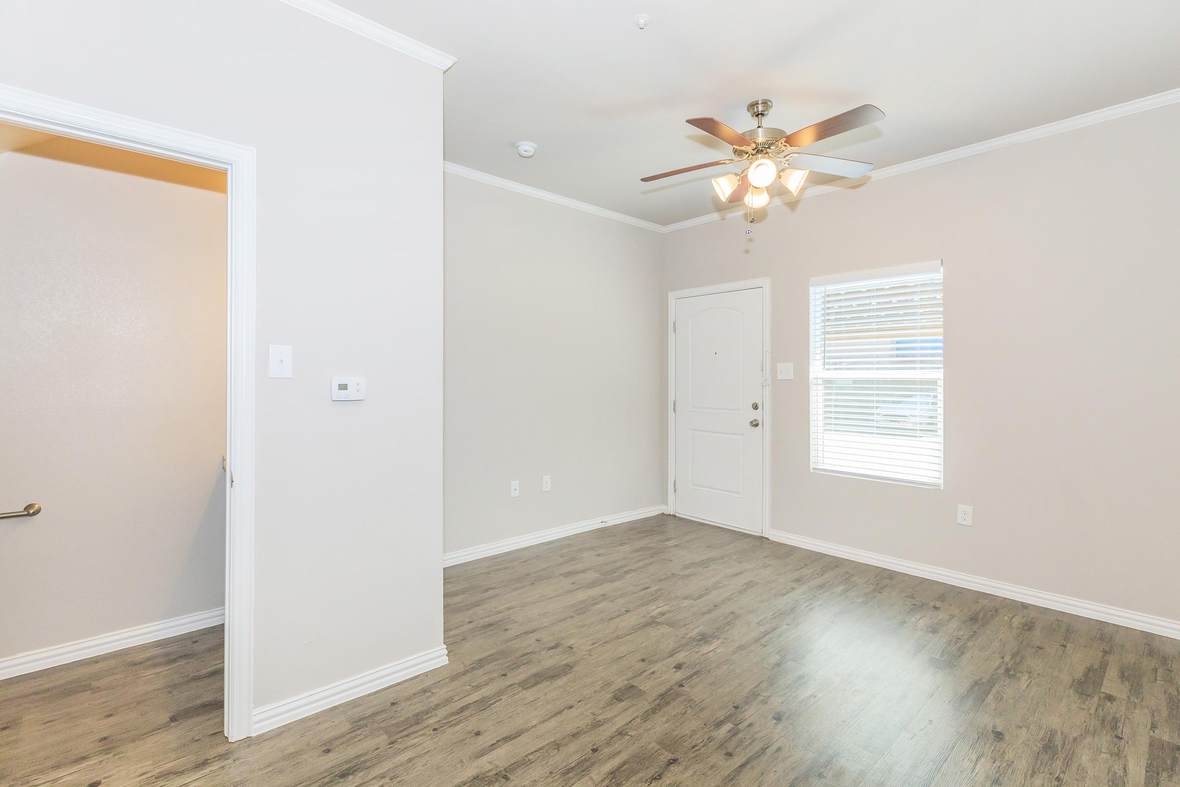A spacious, well-lit room with light gray walls and laminate flooring. It features a ceiling fan, a door leading outside, a window with blinds, and a thermostat on the wall. The room has a simple, modern design and is ready for furniture arrangement.