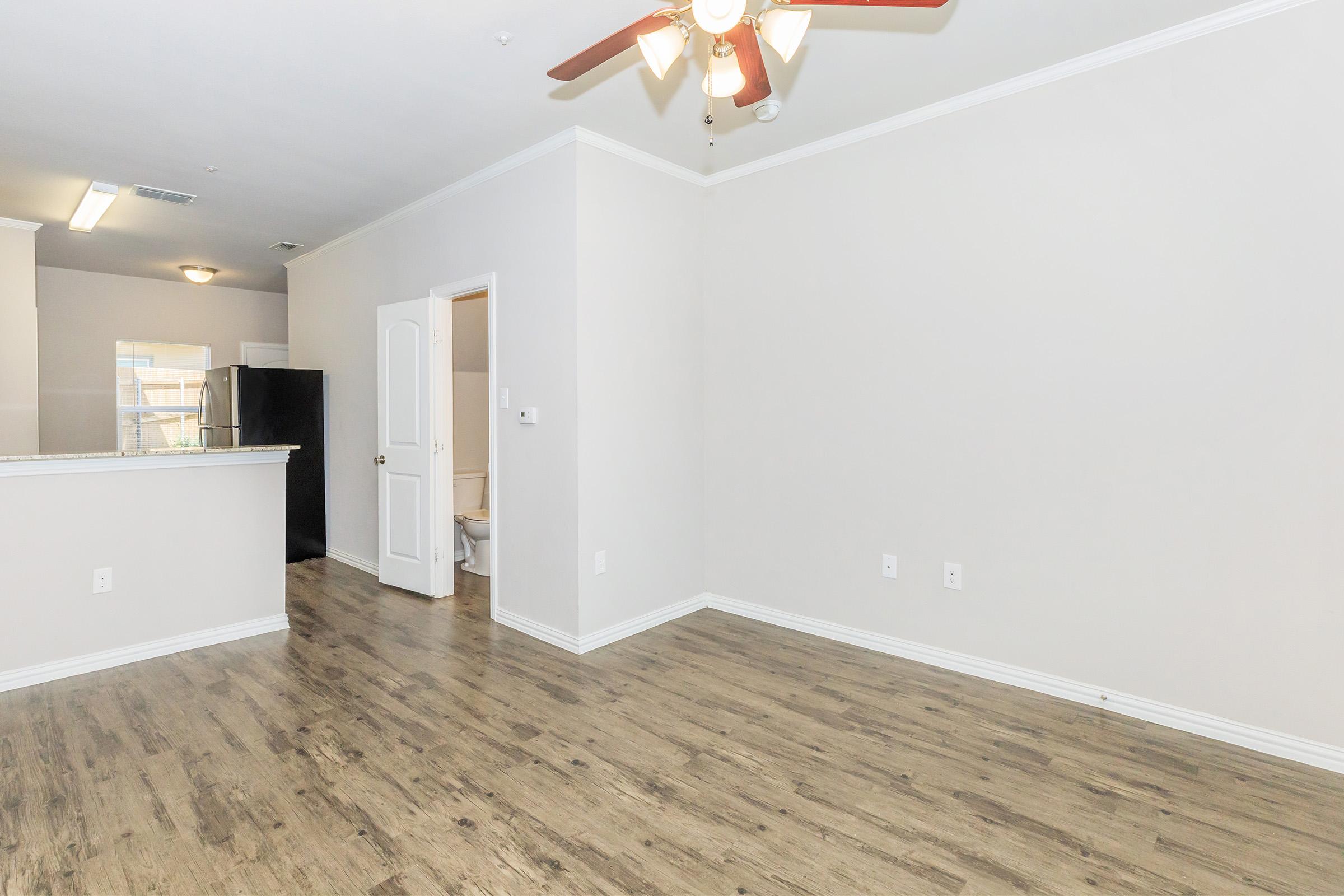 A spacious, empty living area featuring light-colored walls and a ceiling fan. The floor is made of hardwood, and thereβs a doorway leading to a bathroom. To the left, part of a kitchen area is visible, including a refrigerator. The overall ambiance is bright and inviting, suitable for decorating.