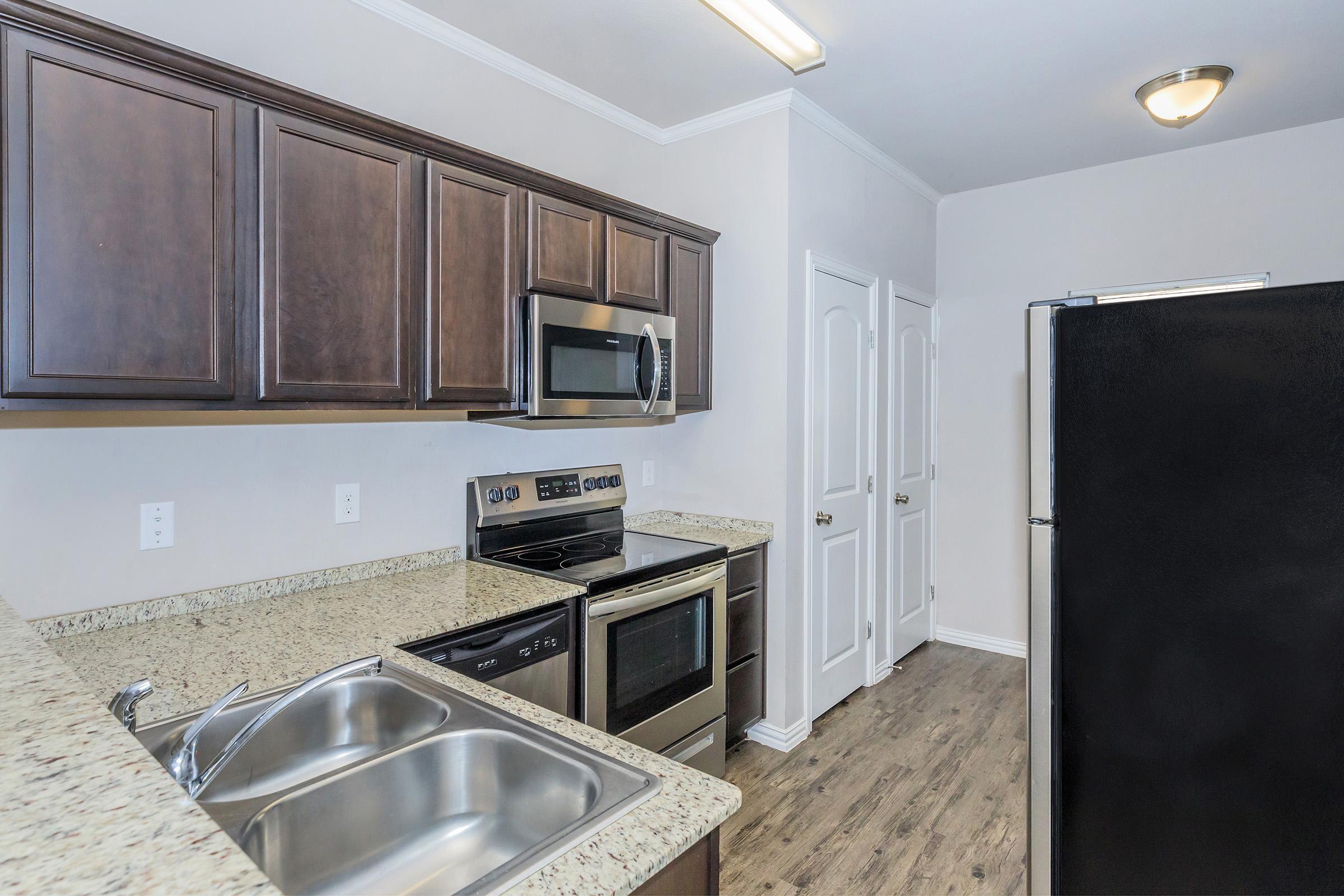 Modern kitchen featuring dark wood cabinets, stainless steel appliances, granite countertops, and a double sink. The design includes an oven, microwave, and refrigerator, with a light-colored wall and wood-look flooring. Two doors lead to additional rooms in the background.