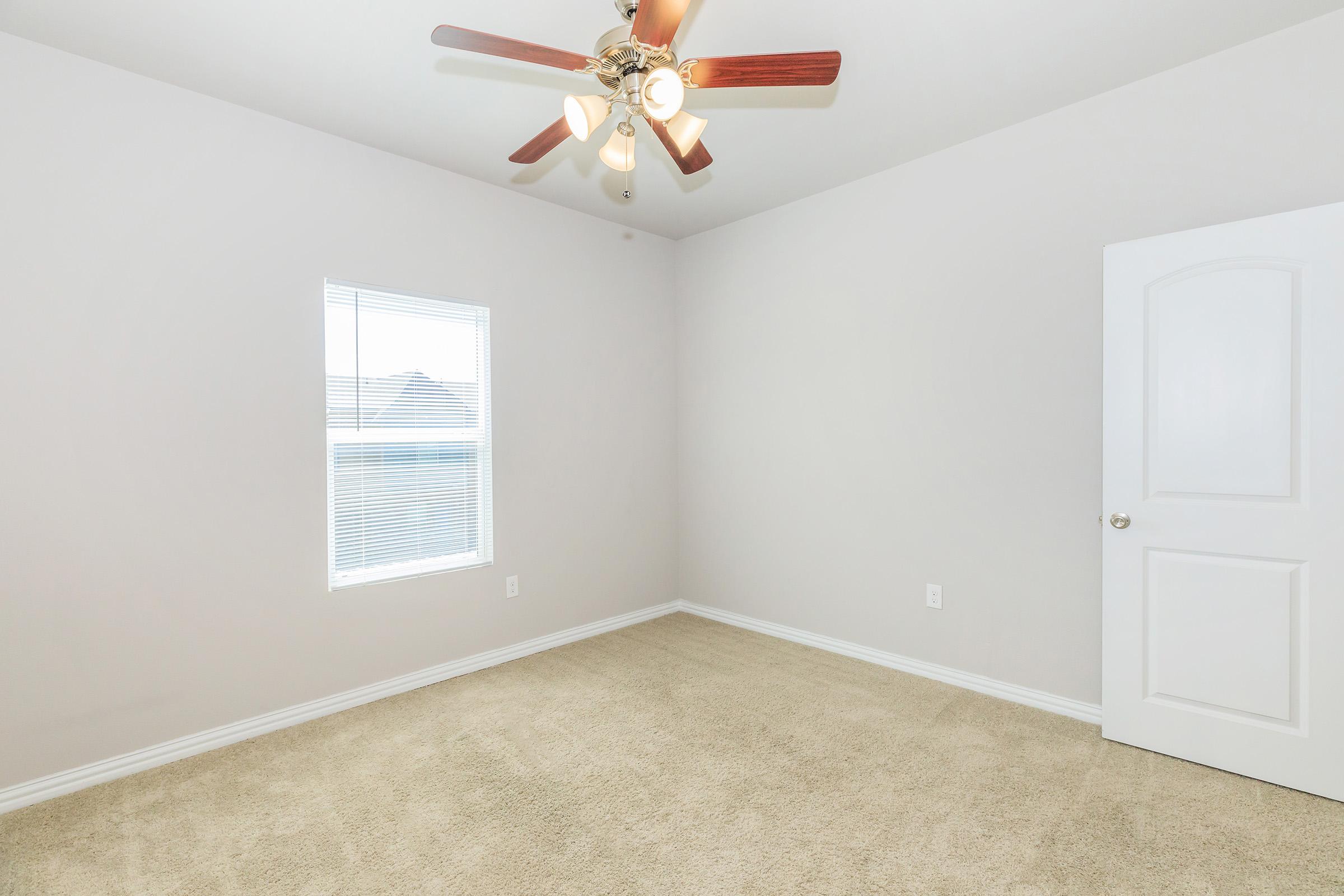 A well-lit, empty room with light beige carpet and gray walls. There's a ceiling fan with wooden blades, and a window allowing natural light. A white door is visible, leading to another space. The overall atmosphere is clean and minimalistic, suitable for various uses.