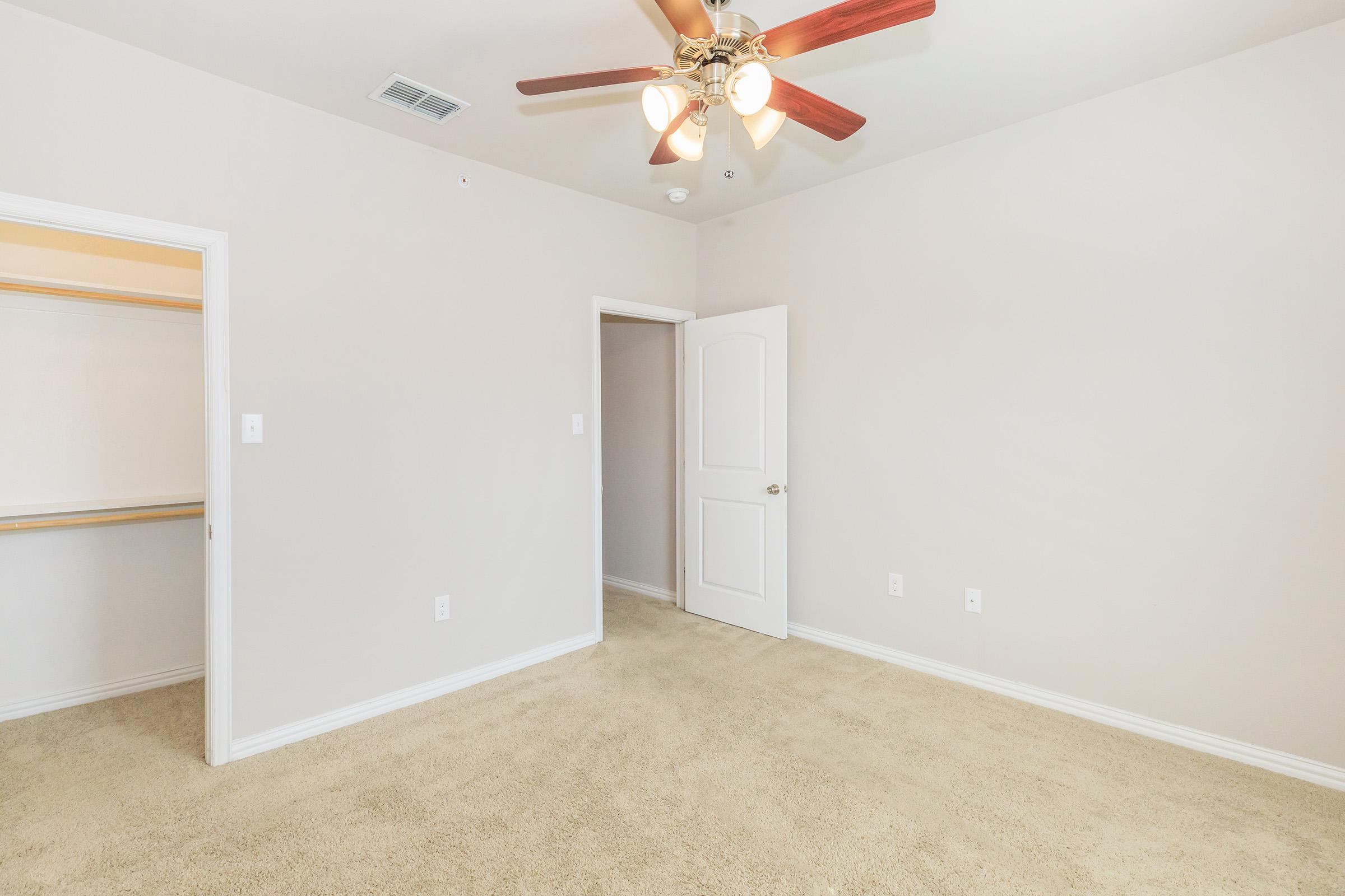 A spacious room with beige walls and a light brown carpet. It features a ceiling fan with wooden blades, a closed closet with a sliding door, and an open door leading to another space. The room is well-lit and devoid of furniture, offering a clean and airy ambiance.