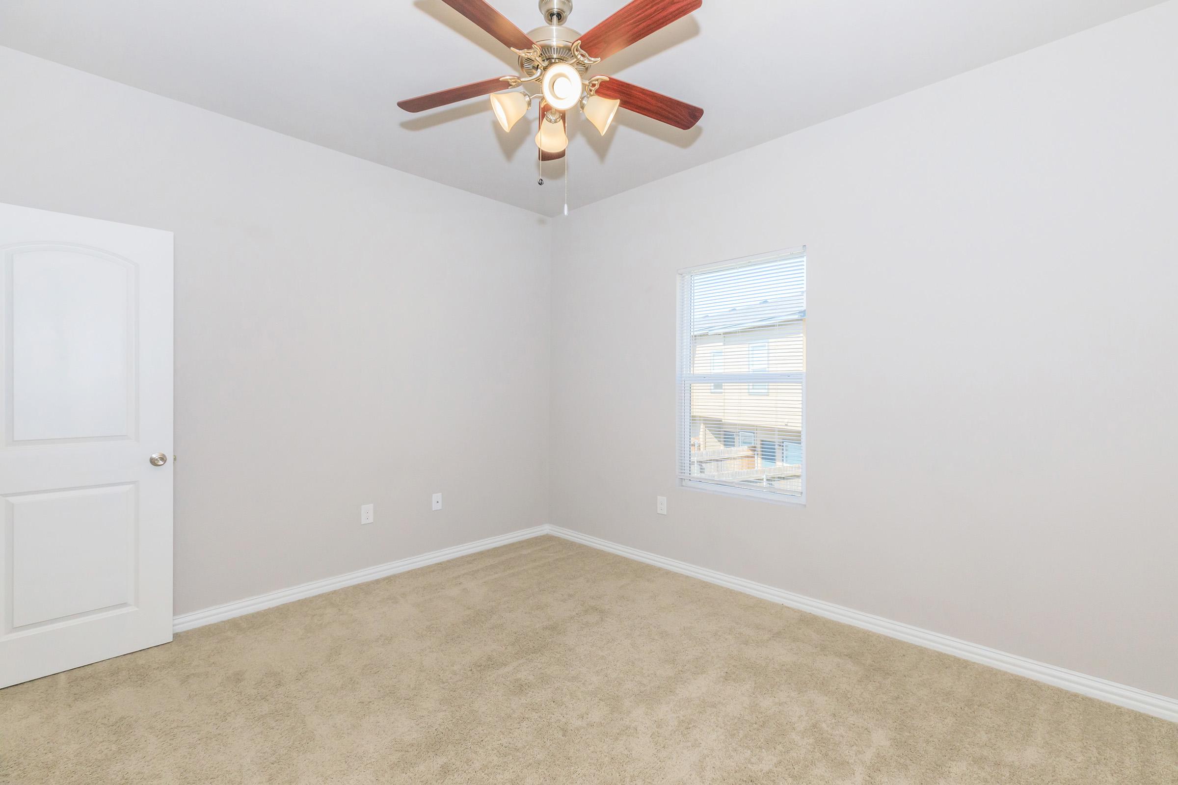 A vacant room with beige carpeting, a light-colored wall, a ceiling fan with wooden blades, and a window allowing natural light. A door is visible on the left side, while the overall space appears empty and ready for furniture.