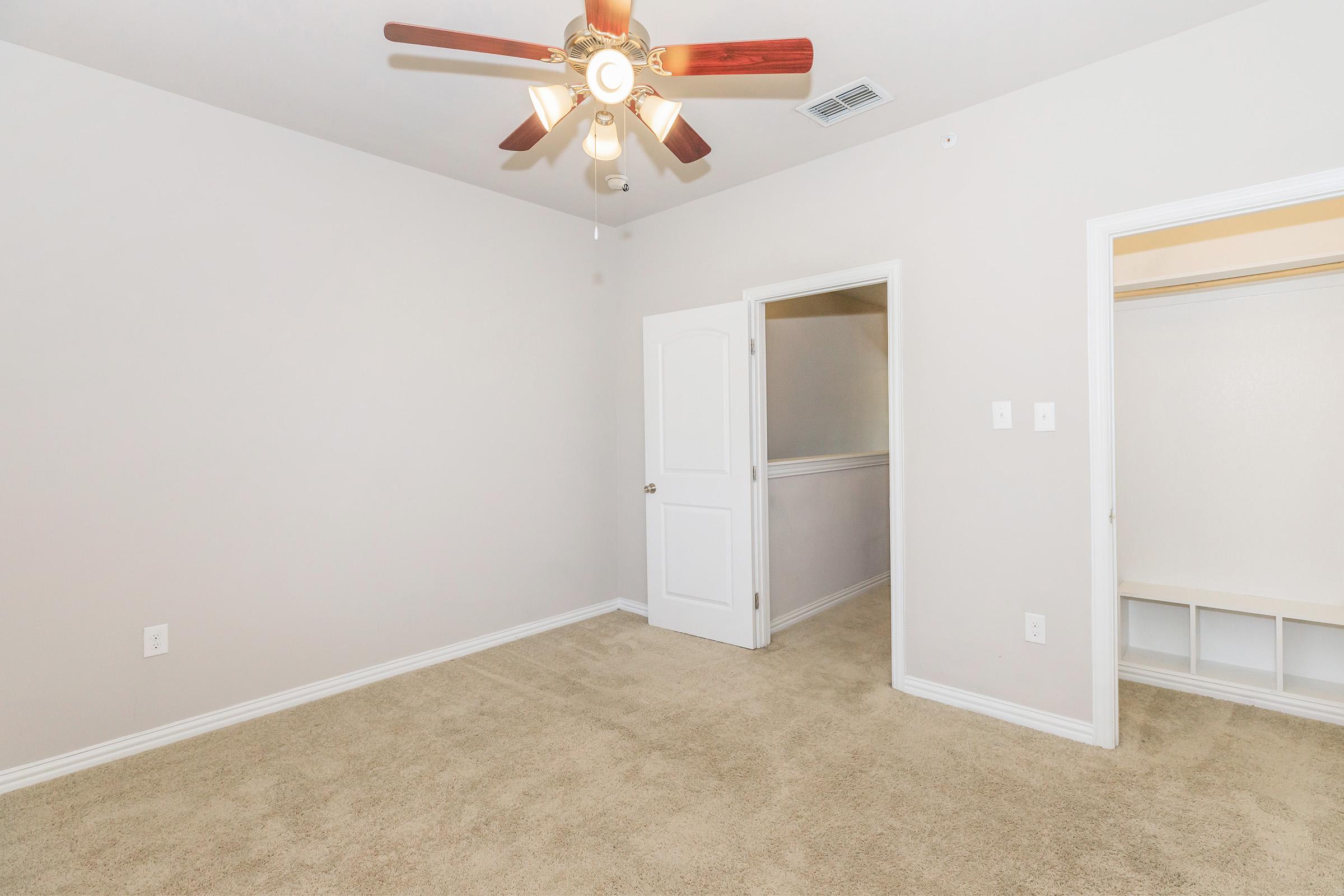 Empty room with light beige walls and carpet. There's a ceiling fan with wooden blades, and doors leading to a closet and another room. The space is well-lit and features a simple, neutral design.