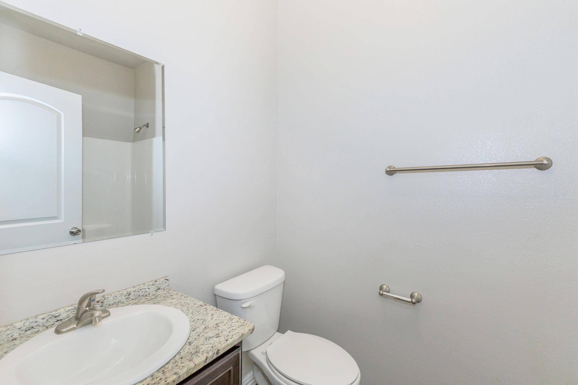 A clean, modern bathroom featuring a white toilet, a granite countertop with a sink, a mirror above the sink, and a towel bar on the wall. The walls are painted a light color, and there is a shower area behind a frosted glass panel.
