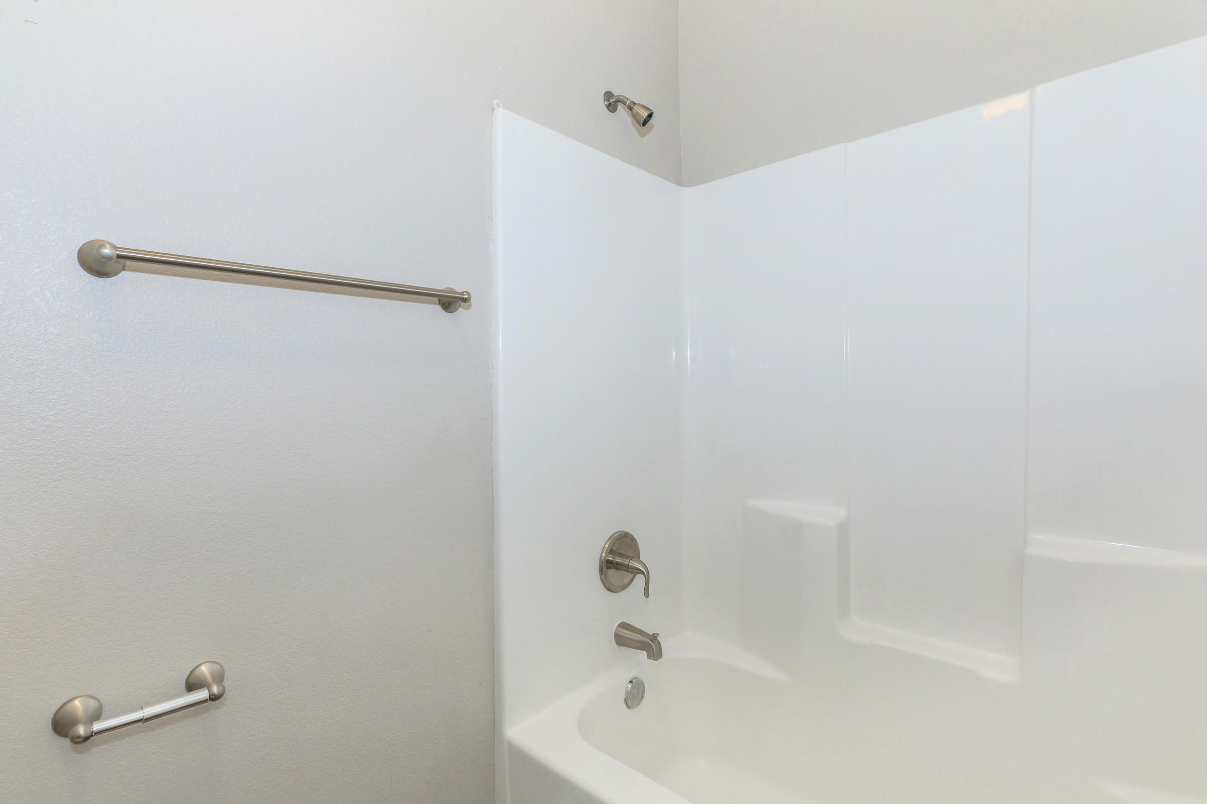 A clean, modern bathroom featuring a white bathtub with a showerhead, chrome fixtures, and a grab bar. The walls are smooth and light-colored, creating a bright and spacious atmosphere.