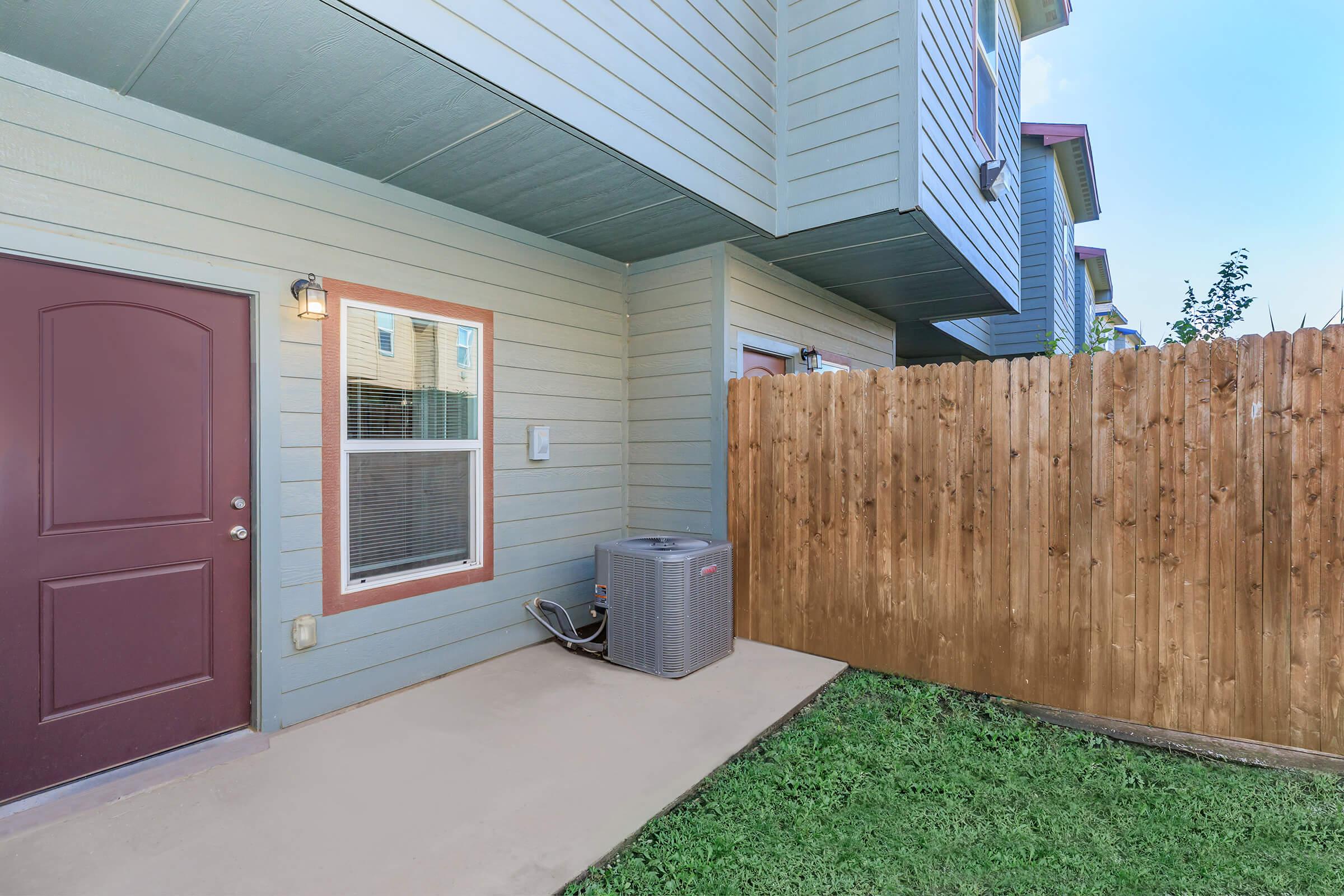 A corner of a home featuring a brown door, a window, and a fenced area with grass. An air conditioning unit is positioned against the house wall next to the door. The exterior is painted in light blue, with a wooden fence providing privacy. Bright daylight illuminates the scene.
