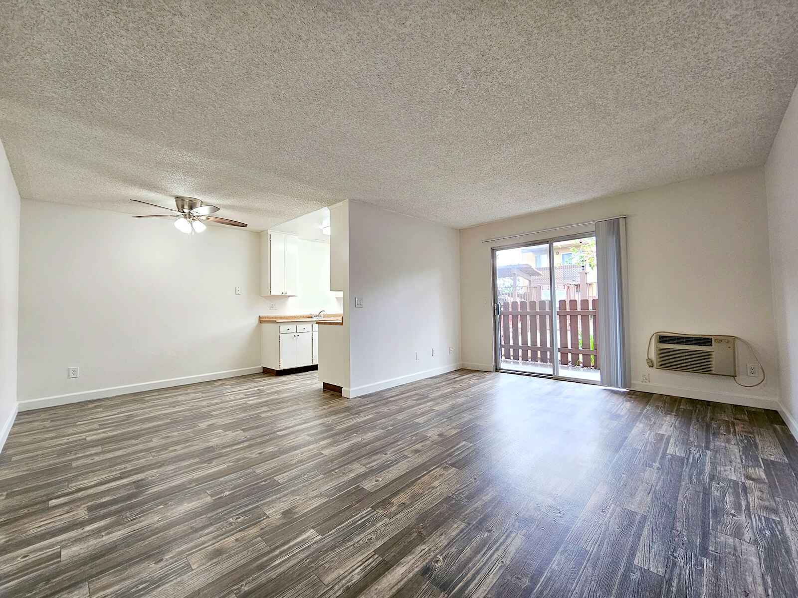 A spacious, empty living room with a ceiling fan, featuring neutral-colored walls and modern laminate flooring. There is a sliding glass door leading to a balcony, and a small kitchen visible in the background with white cabinets. Natural light brightens the space.