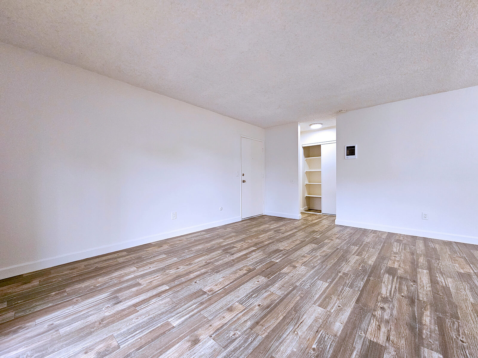 An empty room with light-colored walls and a light wood-like floor. A single door is visible, leading to a hallway. The ceiling is slightly textured, and there is a small wall-mounted item on one side. The space is bright and open, with natural light coming from an unseen window.