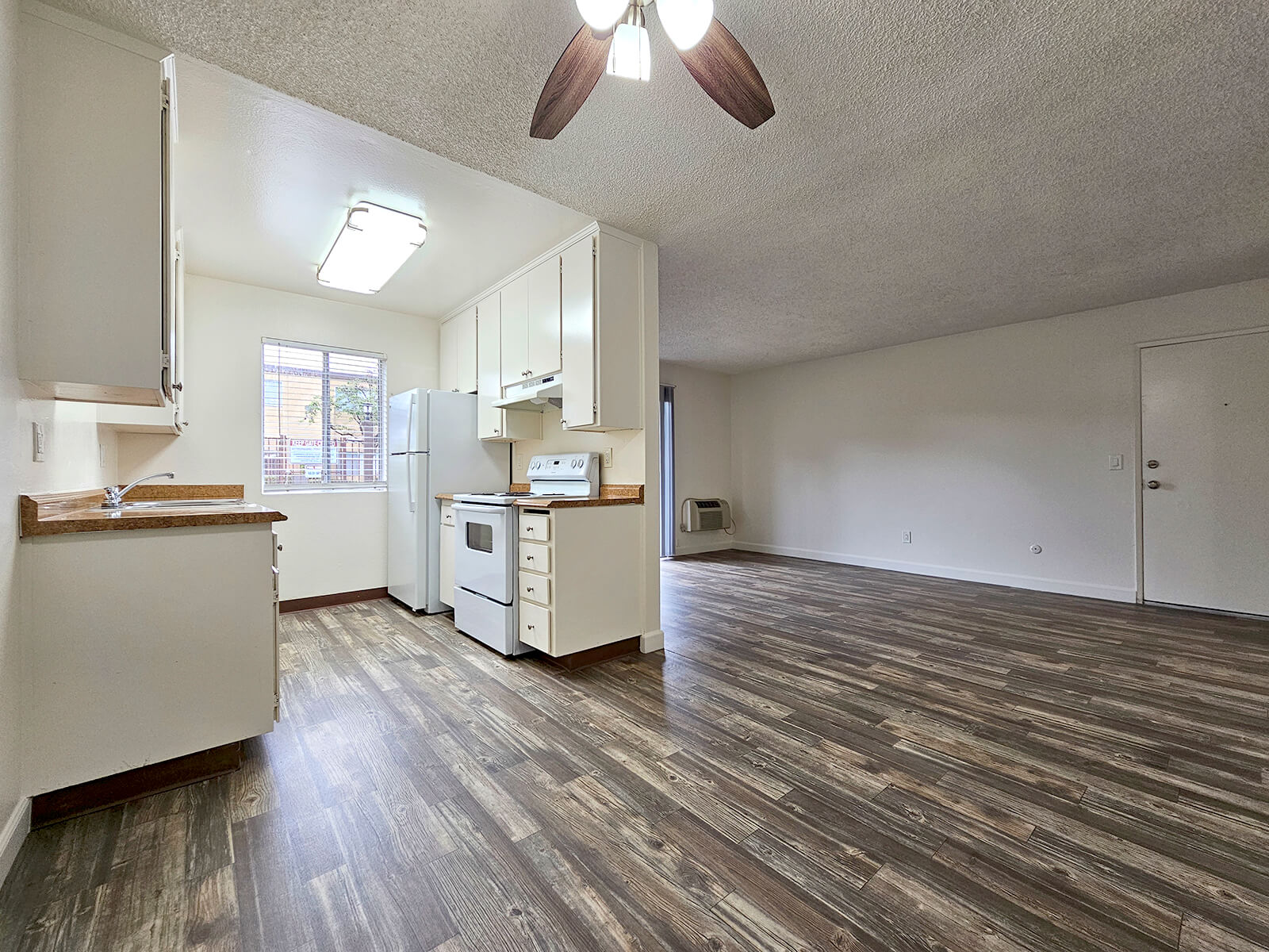 A bright, open-concept kitchen and living area featuring white cabinets, a stove, and a refrigerator. The kitchen is accompanied by a countertop and a window, while the living area has wood-style flooring and a ceiling fan. There is a door leading to an exterior space.