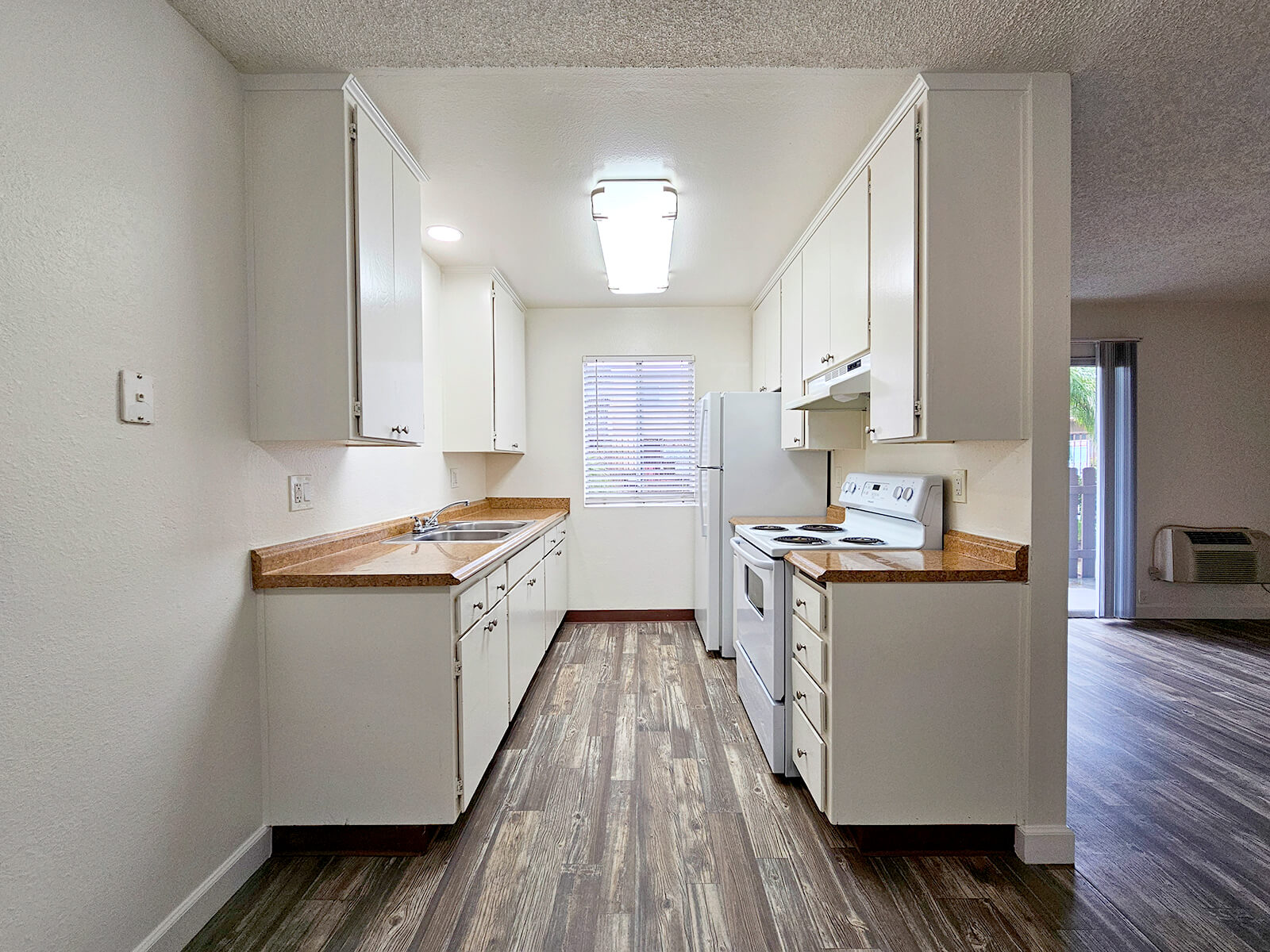 A well-lit kitchen featuring white cabinets, a countertop with a light brown finish, and essential appliances including a stove and refrigerator. The space is clean and modern, with a window providing natural light and wooden flooring enhancing the aesthetic.