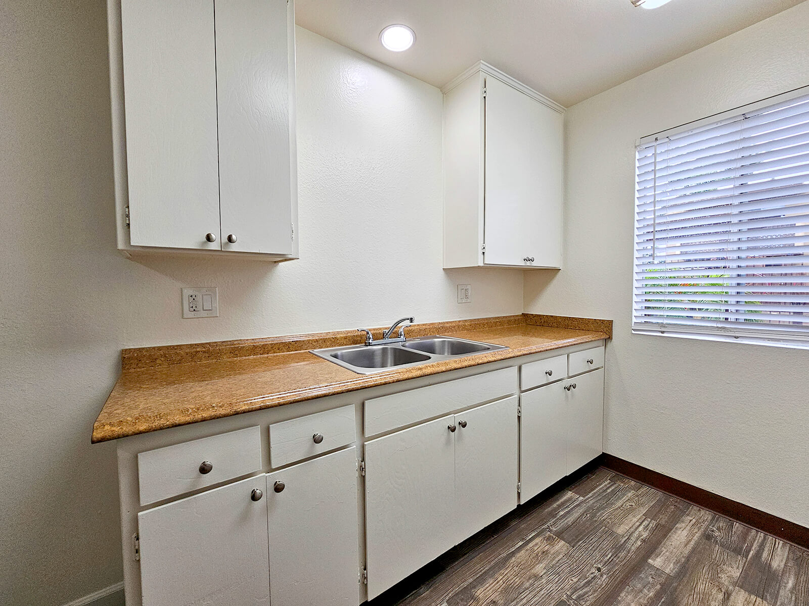 A modern kitchen featuring white cabinets and a light granite countertop. There are two sinks with a faucet in the center, and a window with blinds allowing natural light to enter. The flooring is wooden, adding warmth to the space.