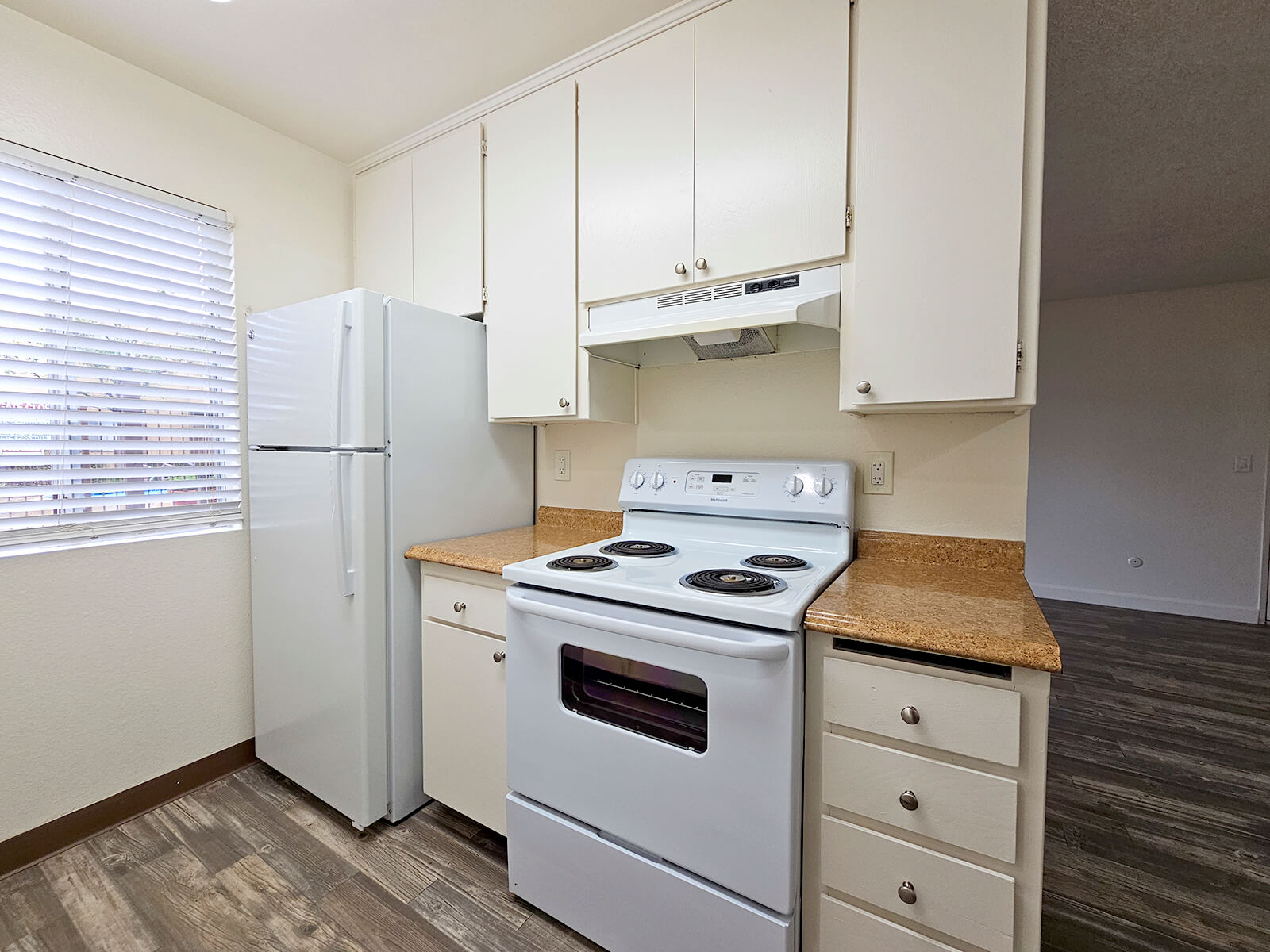 A kitchen with white cabinets, a white refrigerator, and a white stove with an oven. The countertops are light brown, and there is a window with blinds allowing natural light. The flooring is a wood-like design, creating a clean and modern appearance.