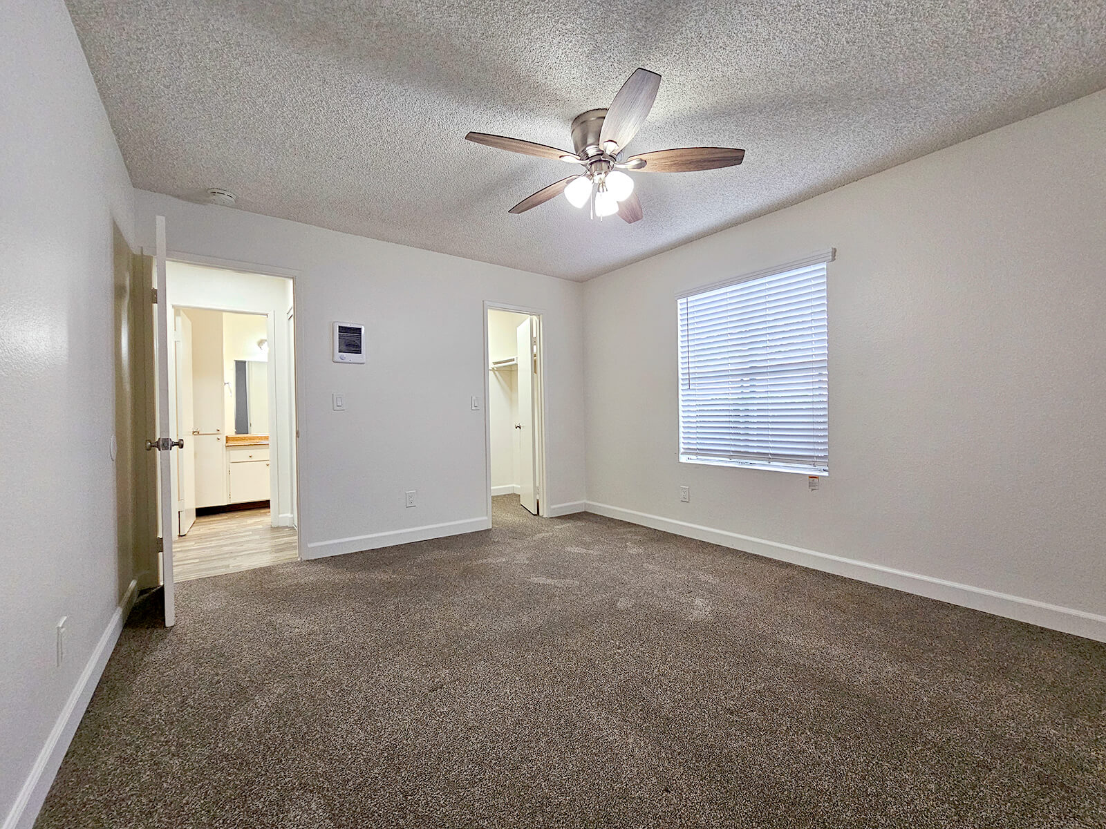 A spacious, well-lit bedroom featuring light-colored walls, a ceiling fan with four blades, and a window with blinds. The room has beige carpeting and a small closet. A doorway leads to a bathroom, and another door opens to a hallway. The overall aesthetic is clean and modern.