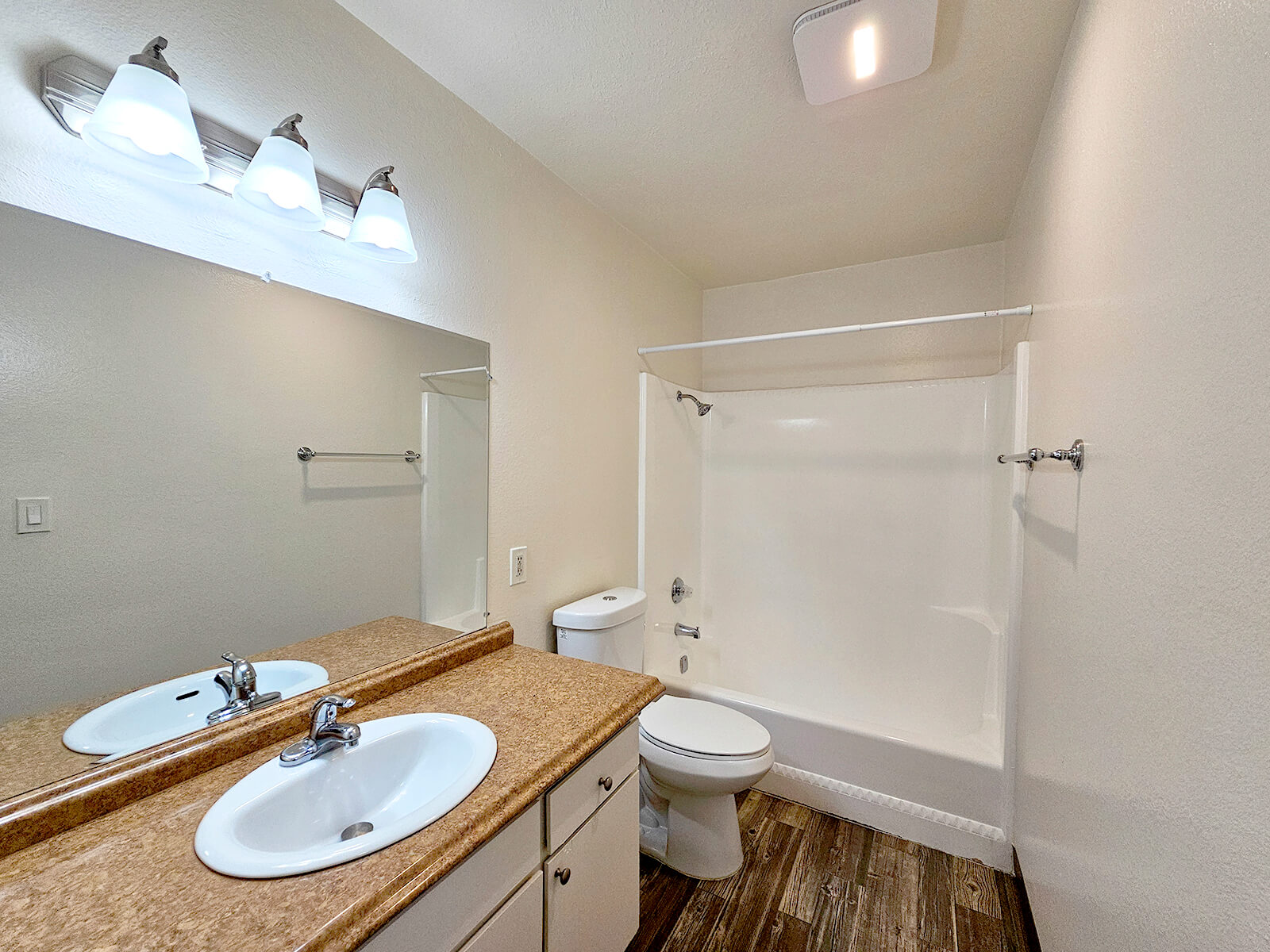 A clean, modern bathroom featuring a double sink vanity with a mirror, a shower/bathtub combo, and a toilet. The walls are painted in a light color, and the flooring has a wood-like appearance. Ceiling lights illuminate the space, creating a bright and airy atmosphere.