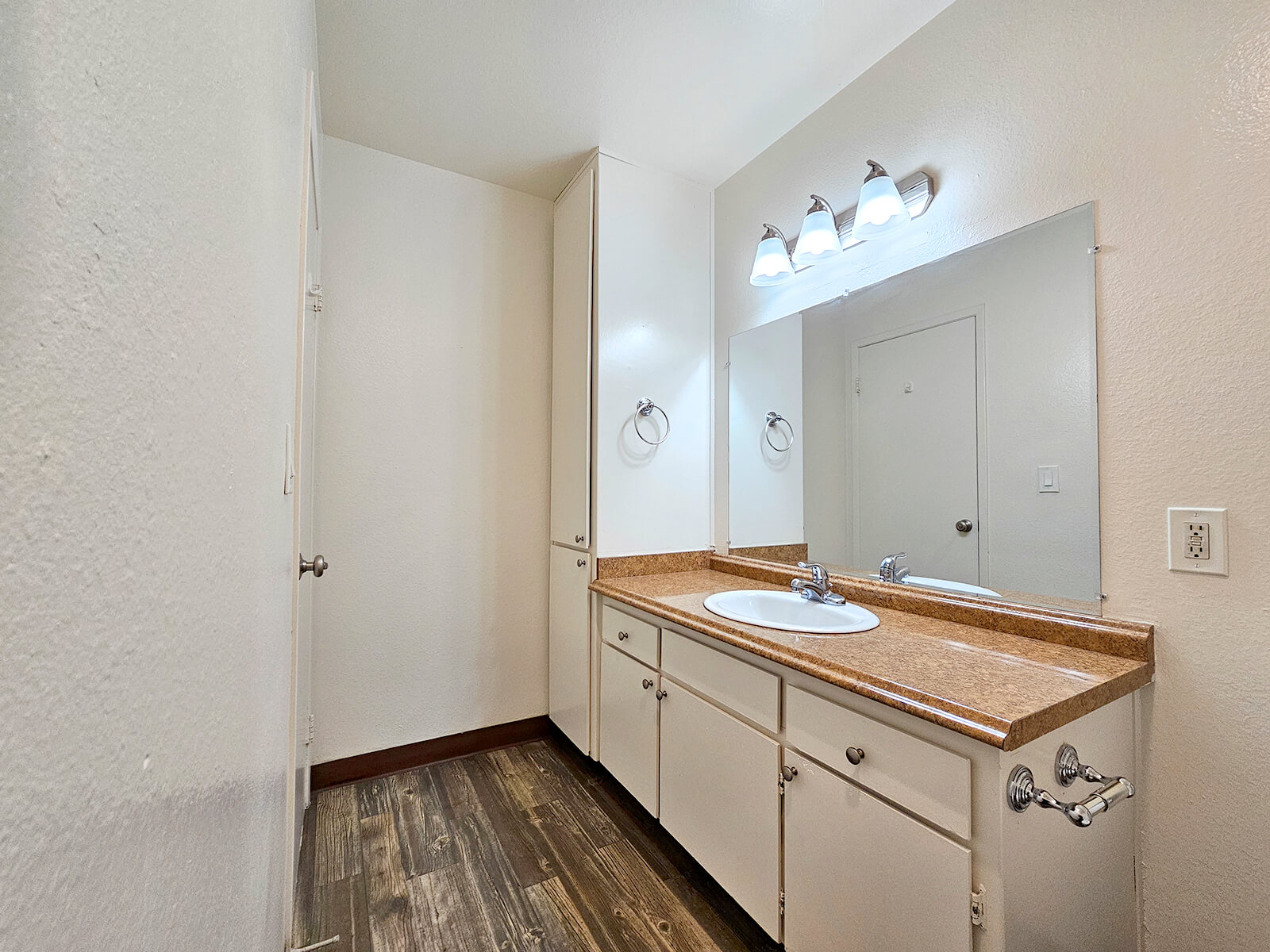 A clean and simple bathroom with a double sink vanity, light-colored walls, wooden flooring, and a large mirror above the sinks. The space includes cabinets for storage and is well-lit with three light fixtures above the mirror. The door to the bathroom is visible on the left.