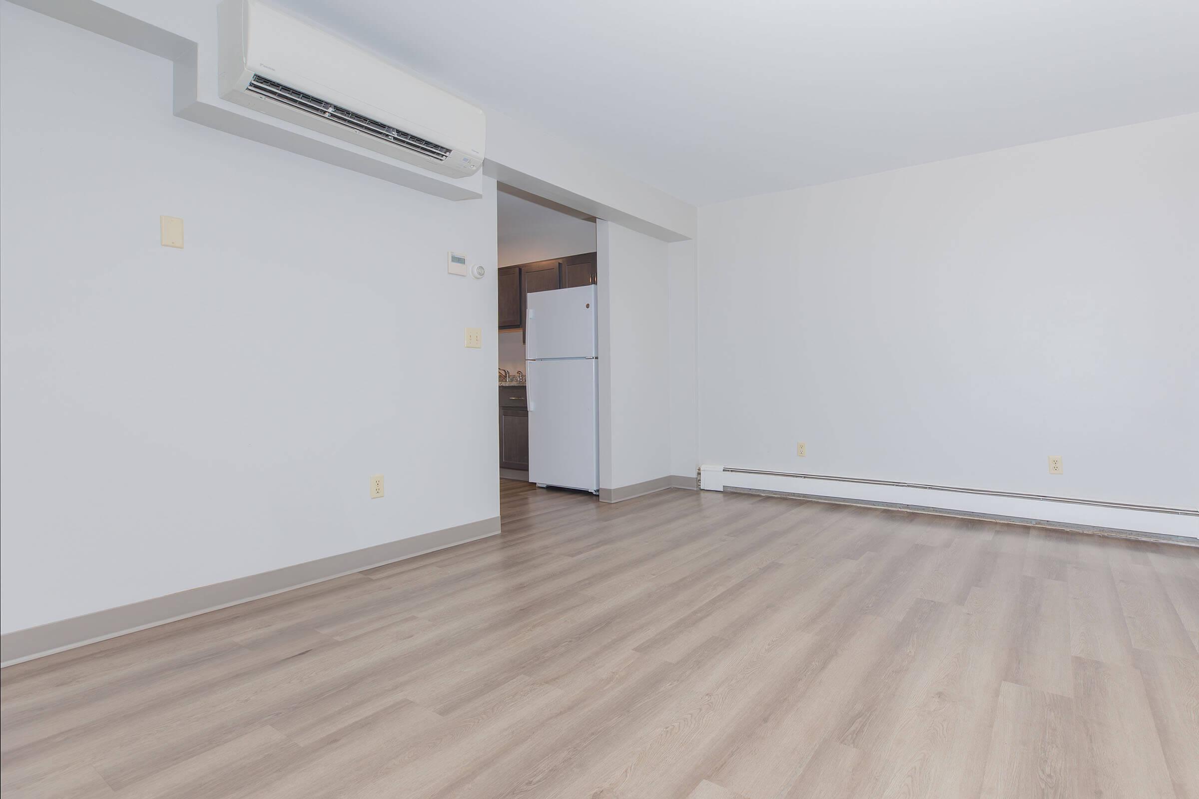 A spacious and empty room with light-colored laminate flooring, featuring a wall-mounted air conditioning unit and a doorway leading to a kitchen area. The walls are painted in a light shade, and there is a refrigerator visible in the background.