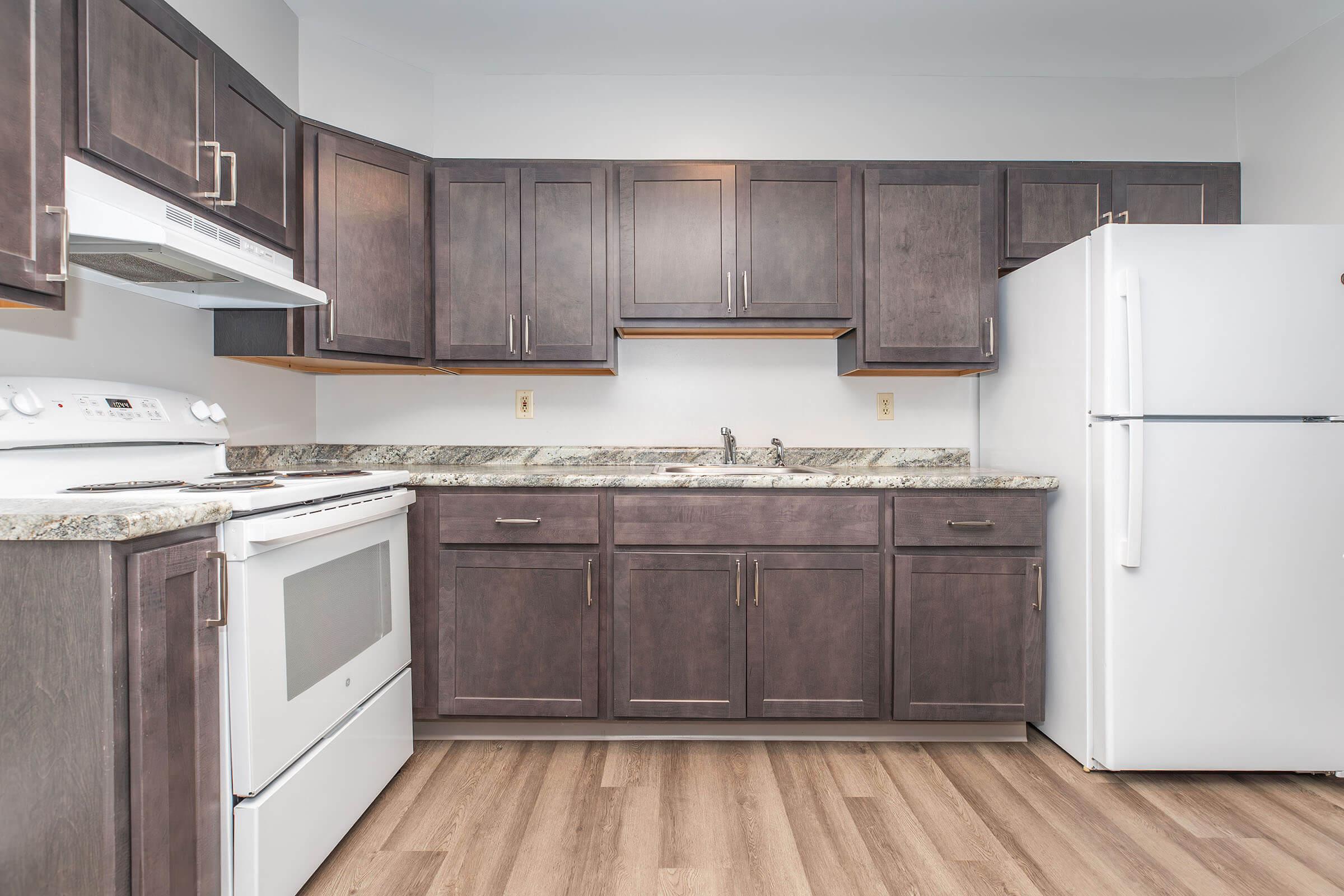 A modern kitchen featuring dark wooden cabinets, a granite countertop, a white stove, and a white refrigerator. There's a sink with a faucet, and the floor is covered in wood-like laminate. The overall design is sleek and contemporary, with natural light illuminating the space.