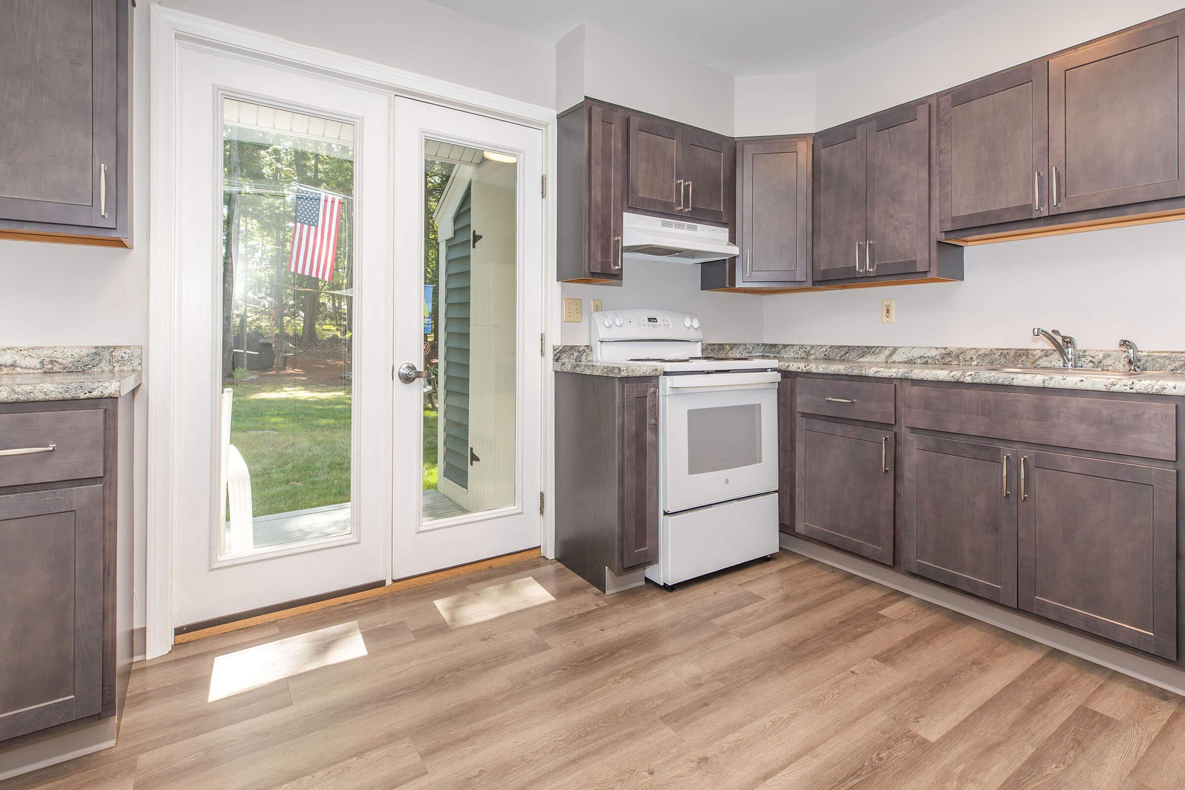 Modern kitchen with dark wood cabinets, a white stove, and a granite countertop. A door with glass panels leads to a backyard, featuring a view of an American flag outside. The flooring is light-colored wood, creating a bright and inviting atmosphere.
