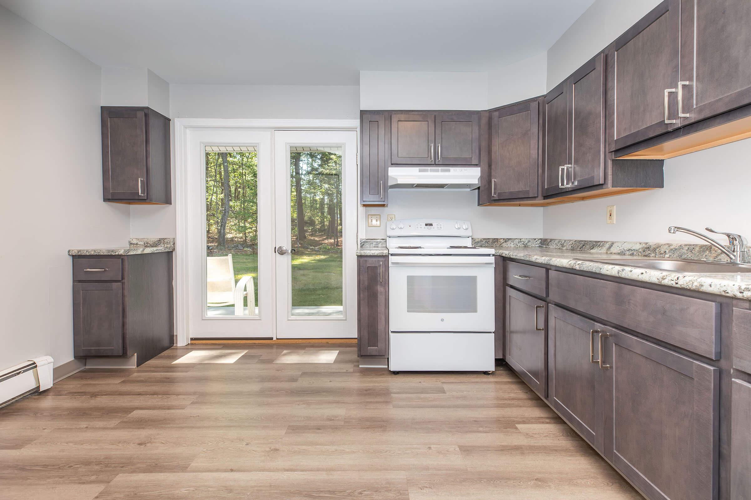 Modern kitchen featuring dark wood cabinets, a white stove, and a granite countertop. There is a sliding glass door leading to an outdoor patio, allowing natural light to fill the space. The floor is finished with a light wood laminate, creating a warm and inviting atmosphere.