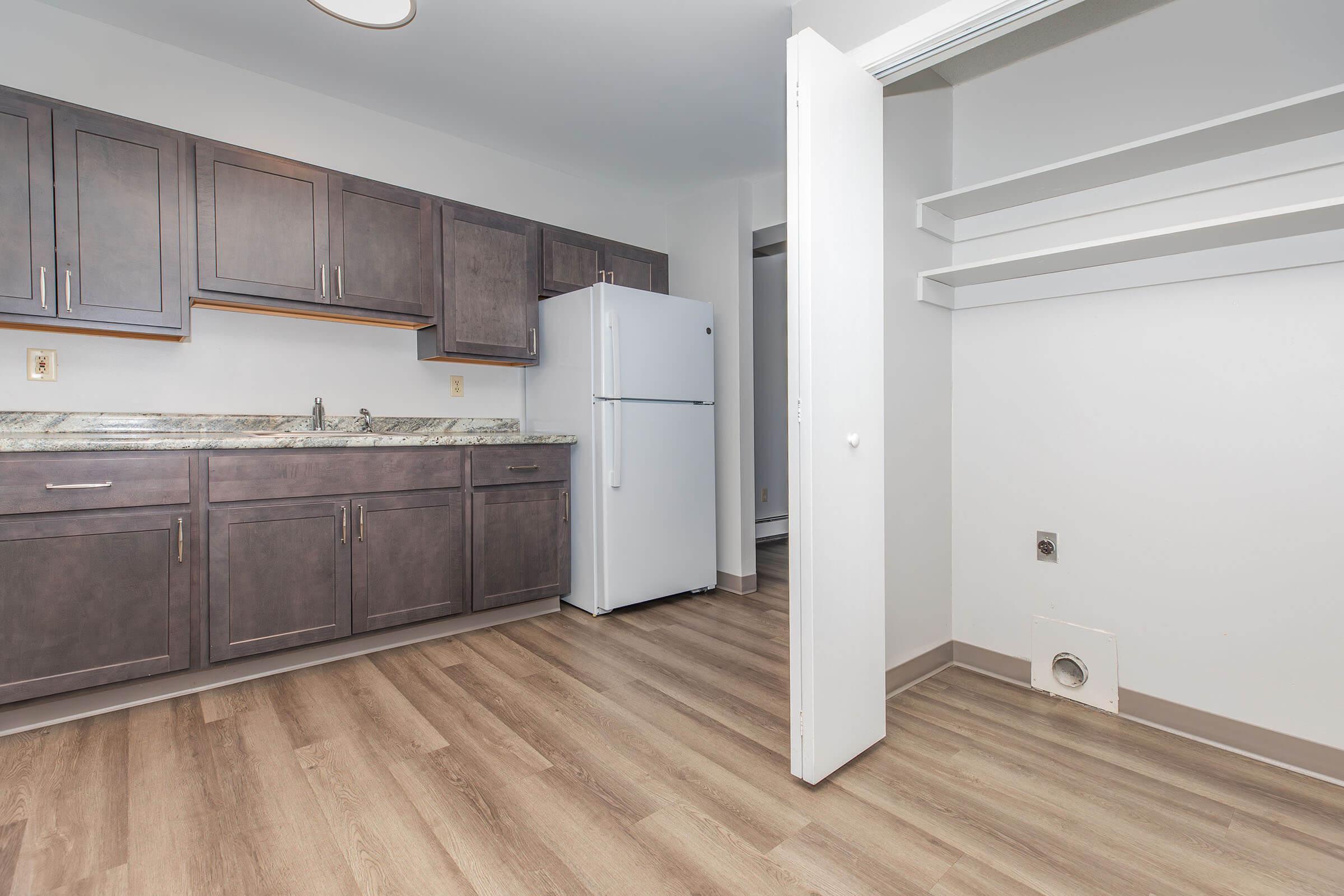 A modern kitchen featuring dark cabinetry, a white refrigerator, and a countertop with a granite finish. The space has a light wood floor, with open shelving on one side and a closet area with a door on the right. The walls are painted in a light color, creating a bright and inviting atmosphere.