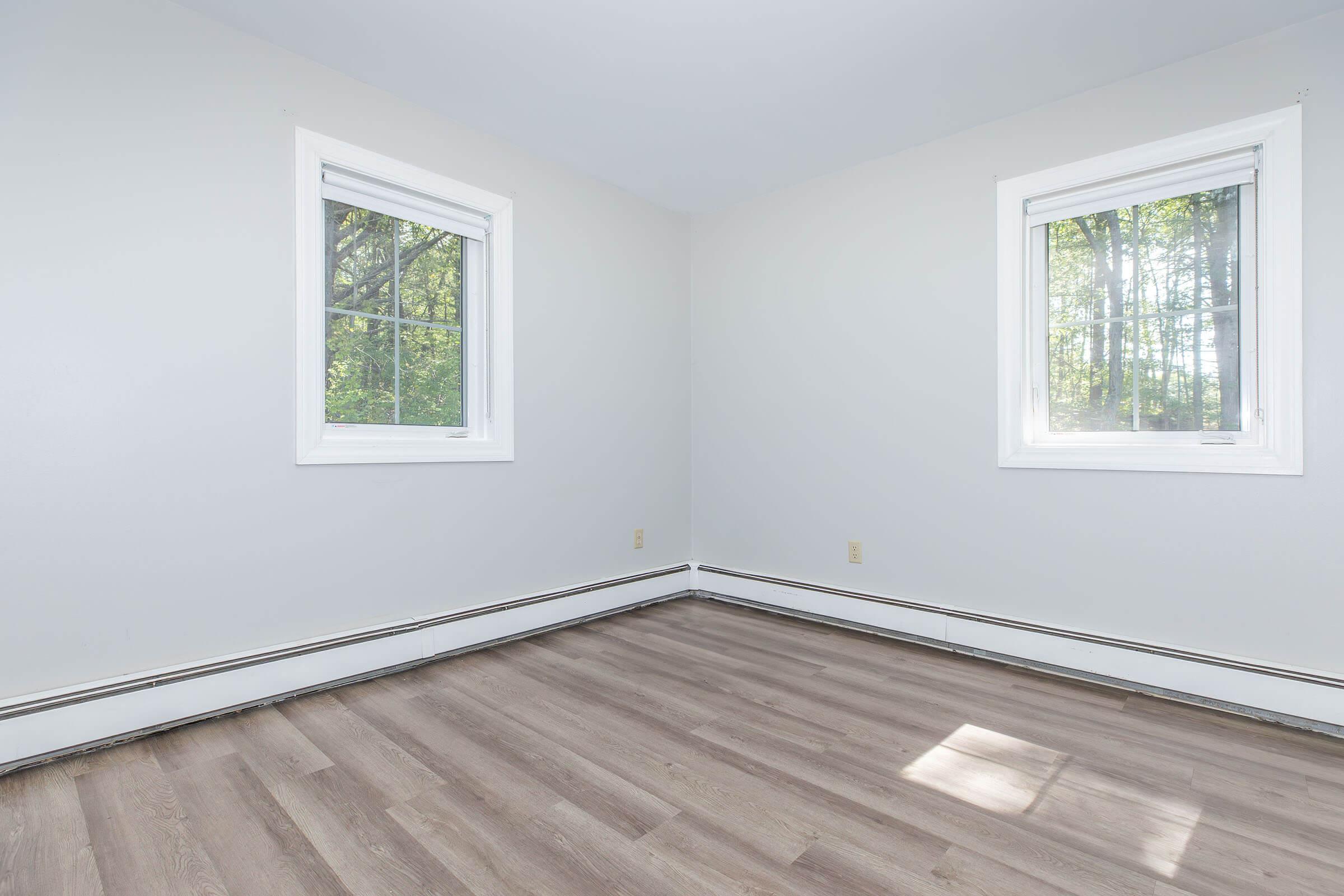 Empty room with light gray walls and two windows allowing natural light. The flooring is light brown, resembling wood. There is no furniture present, creating a spacious and uncluttered atmosphere.