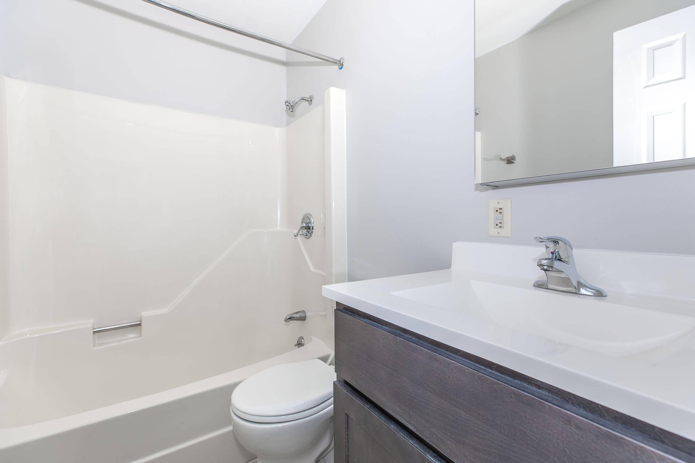 A modern bathroom featuring a white bathtub and shower combination, a sleek countertop with a sink and faucet, and a large mirror above the sink. The walls are painted in a light gray color, and a white toilet is also visible. The overall design is clean and minimalist.